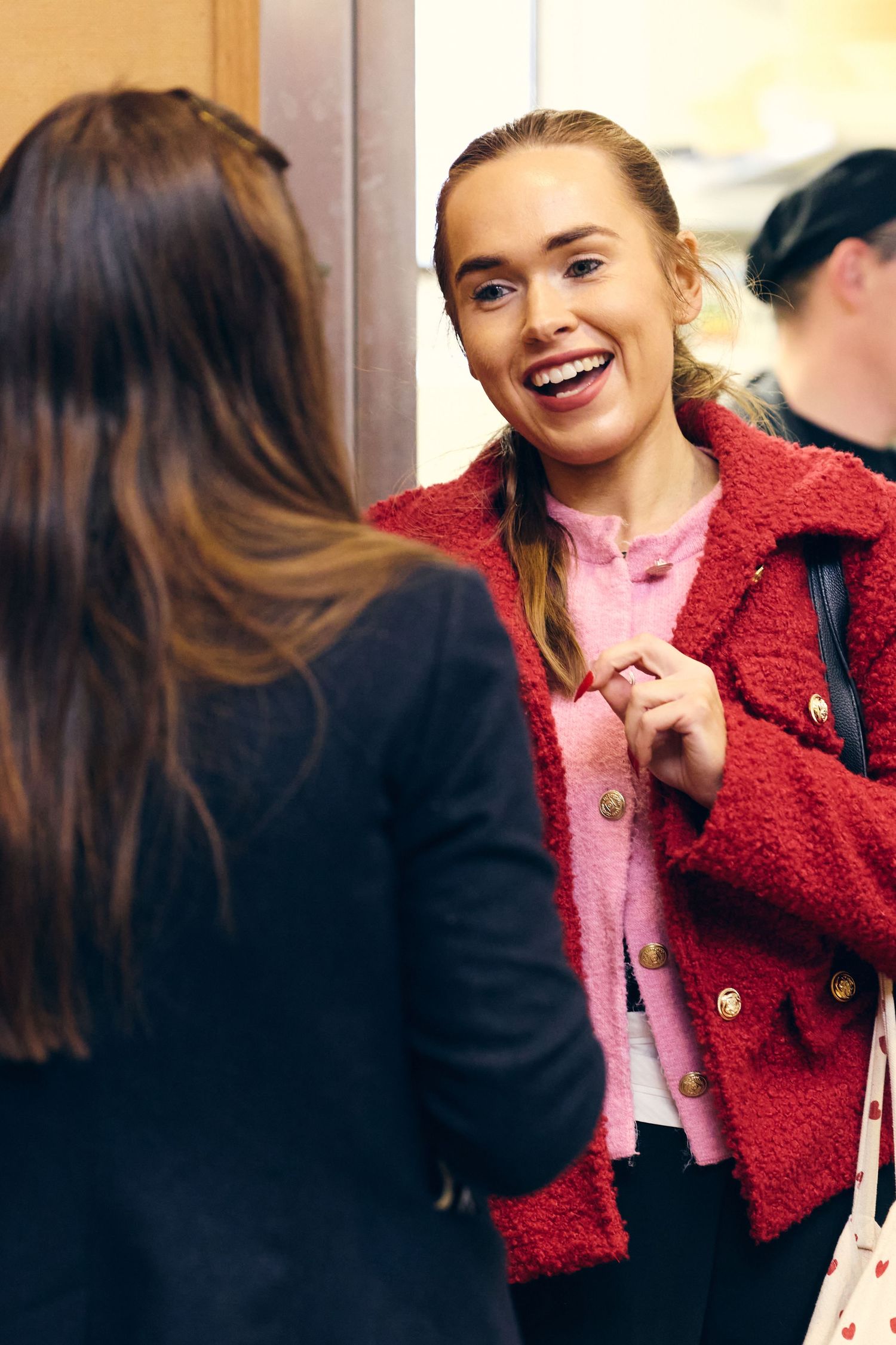 A smiling blonde woman in a red coat chats with a dark-haired woman indoors at a social gathering.