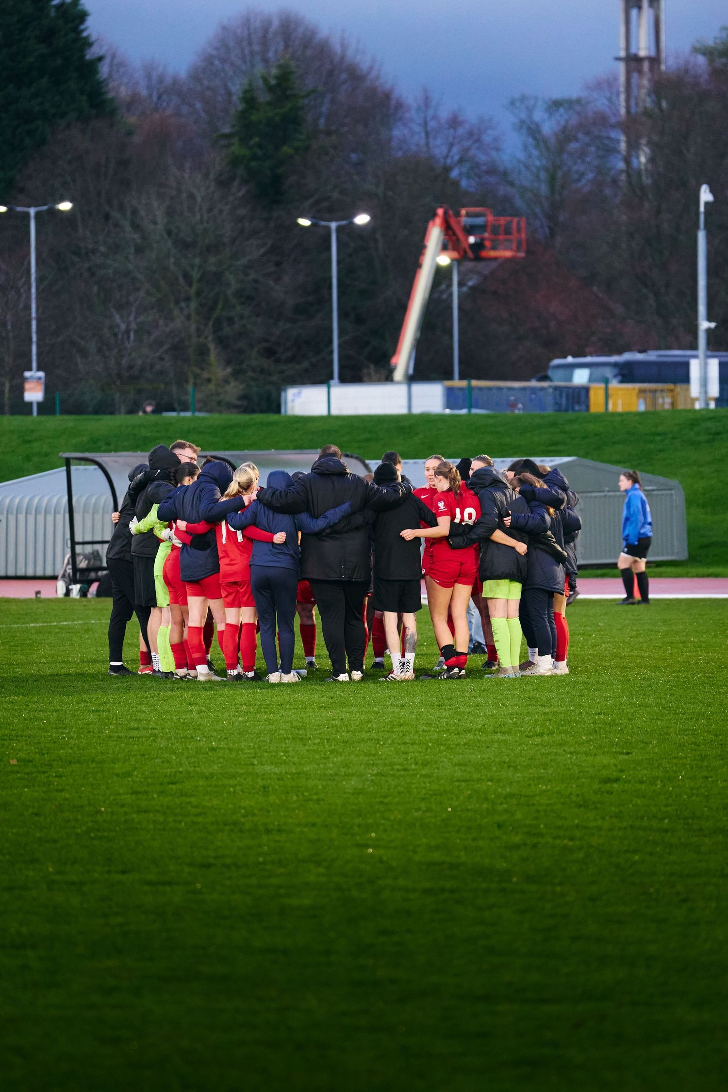 Soccer team huddles together in a tight circle on a grass field during dusk with floodlights illuminating the scene.