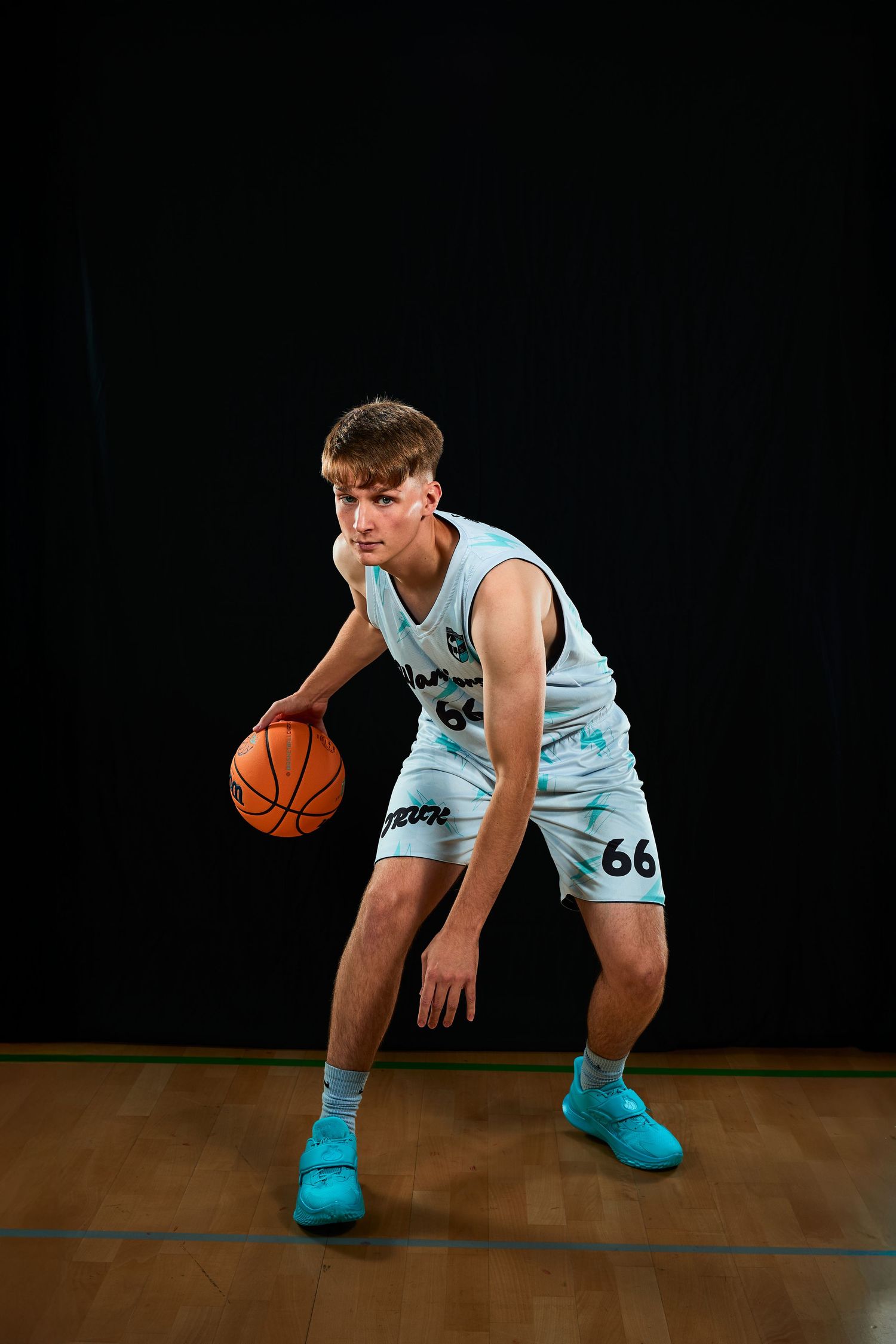Basketball player in light blue jersey number 66 dribbling on wooden court against black background.