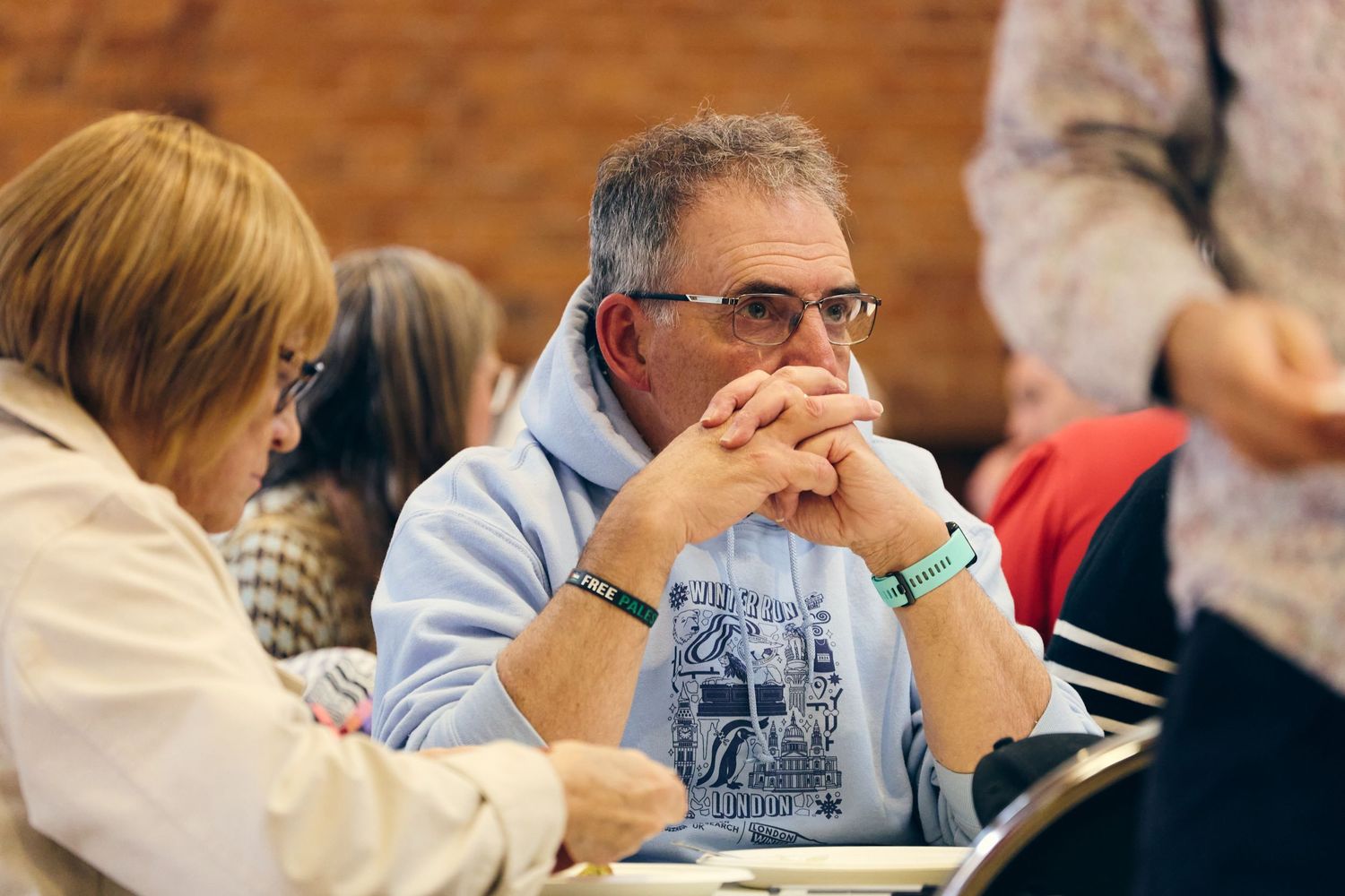 Man with glasses and blue hoodie sits thoughtfully at a table during a seminar or conference event.