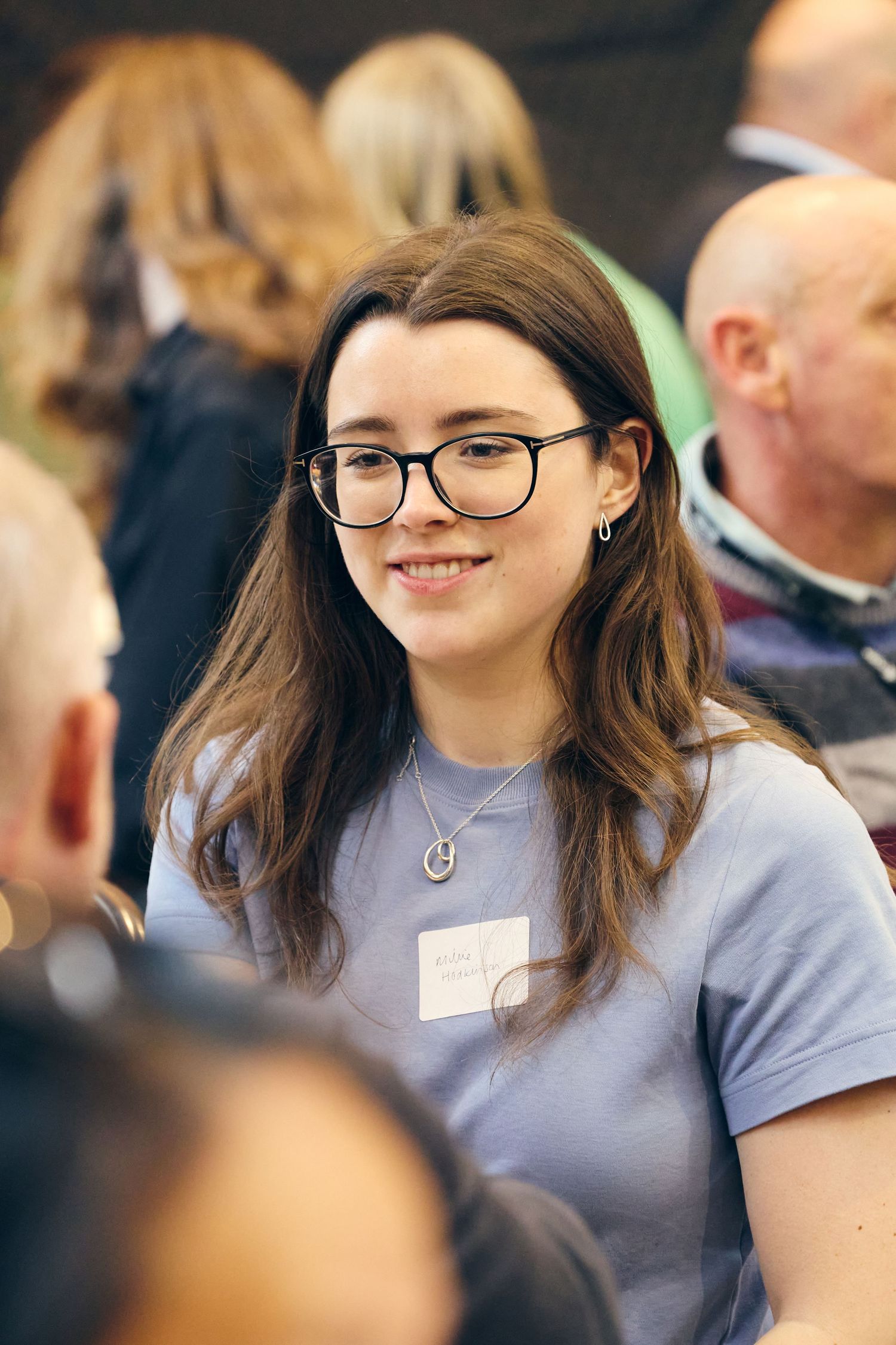 Young woman with glasses and name tag smiling at a networking event, wearing a light blue t-shirt.