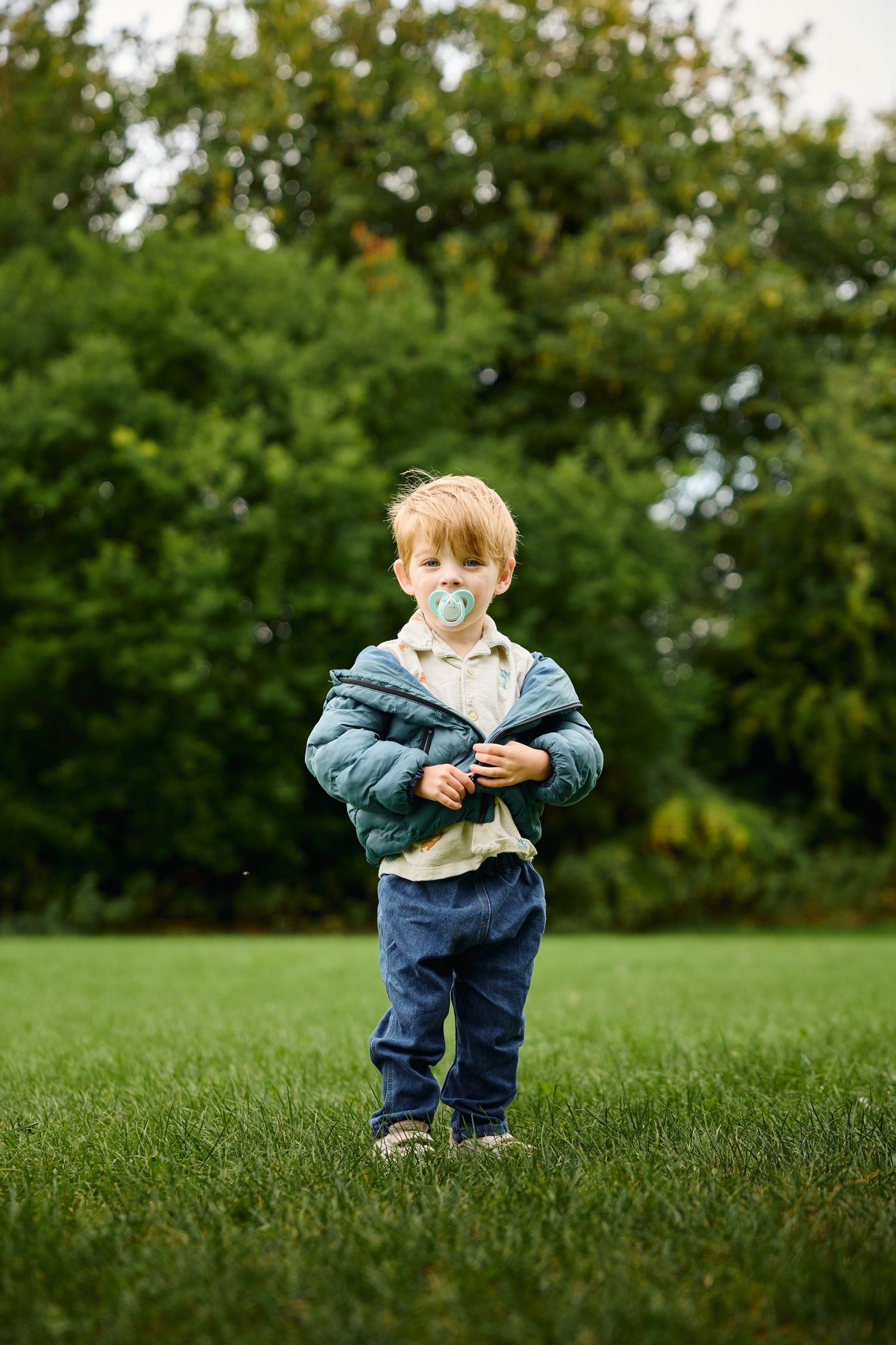 Small child in denim and jacket exploring grassy field with trees in background.