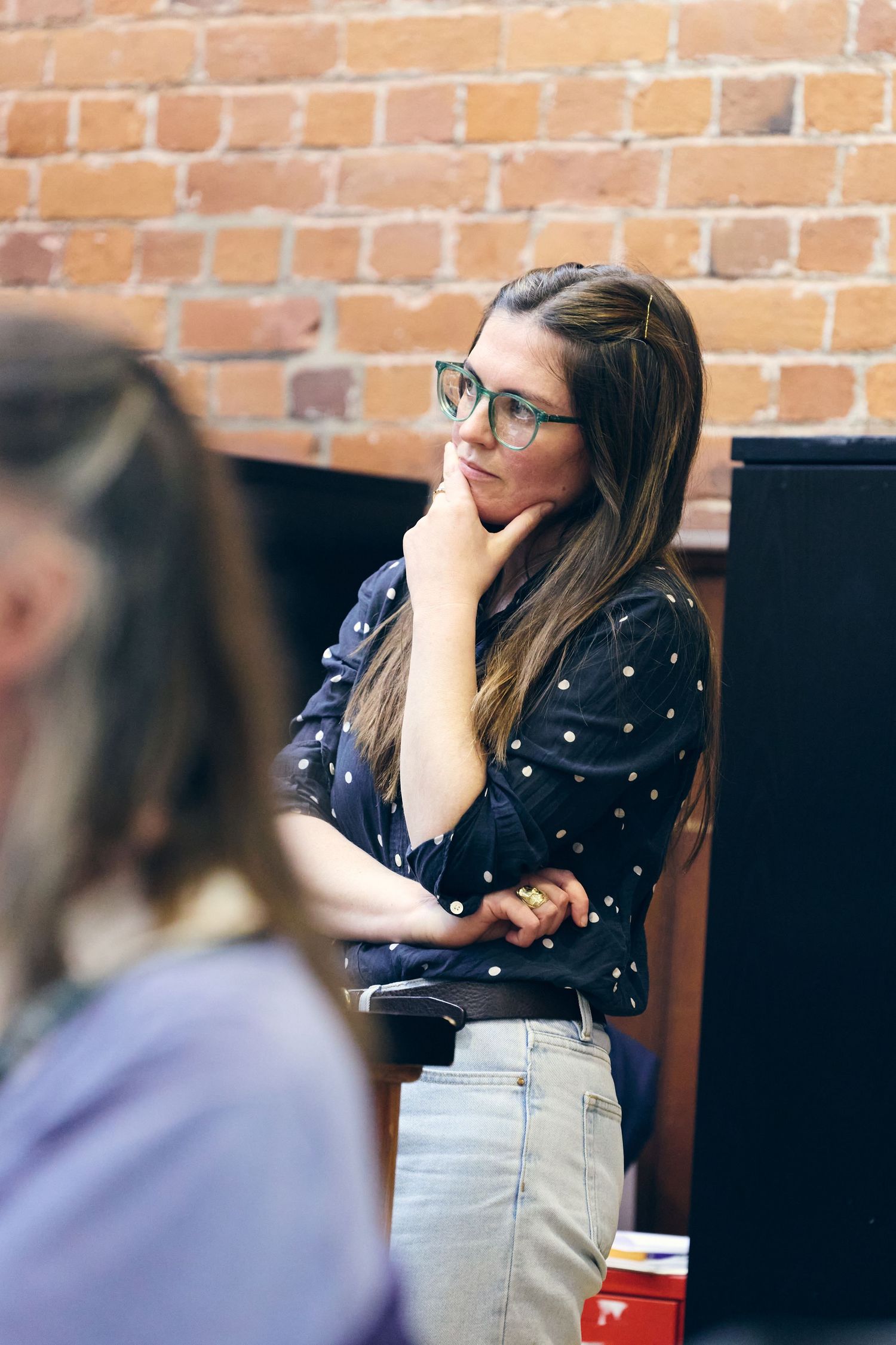 Thoughtful young woman with glasses and polka dot blouse standing in modern office with brick wall.