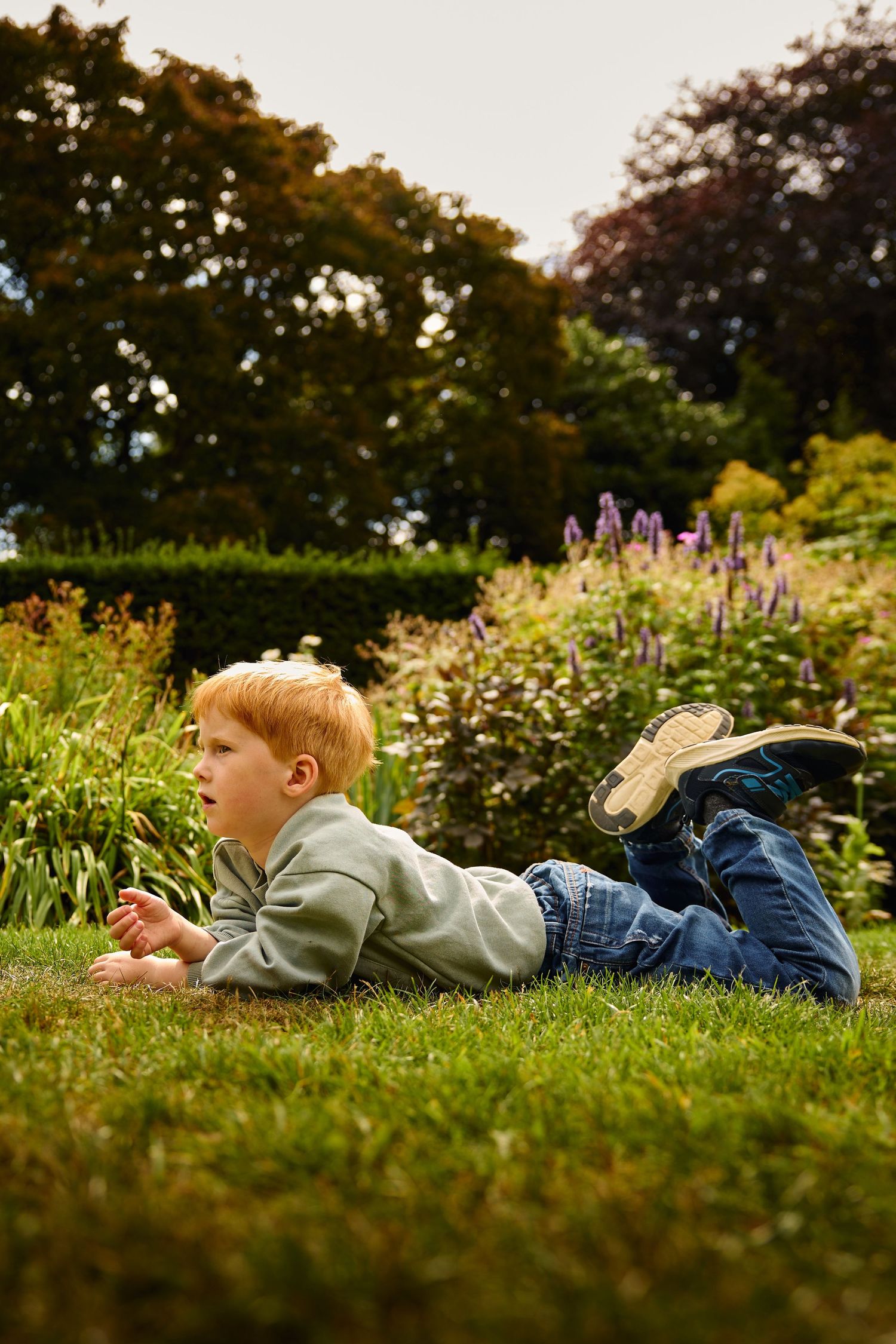 Child lying in grass with colorful garden flowers in background during golden hour.