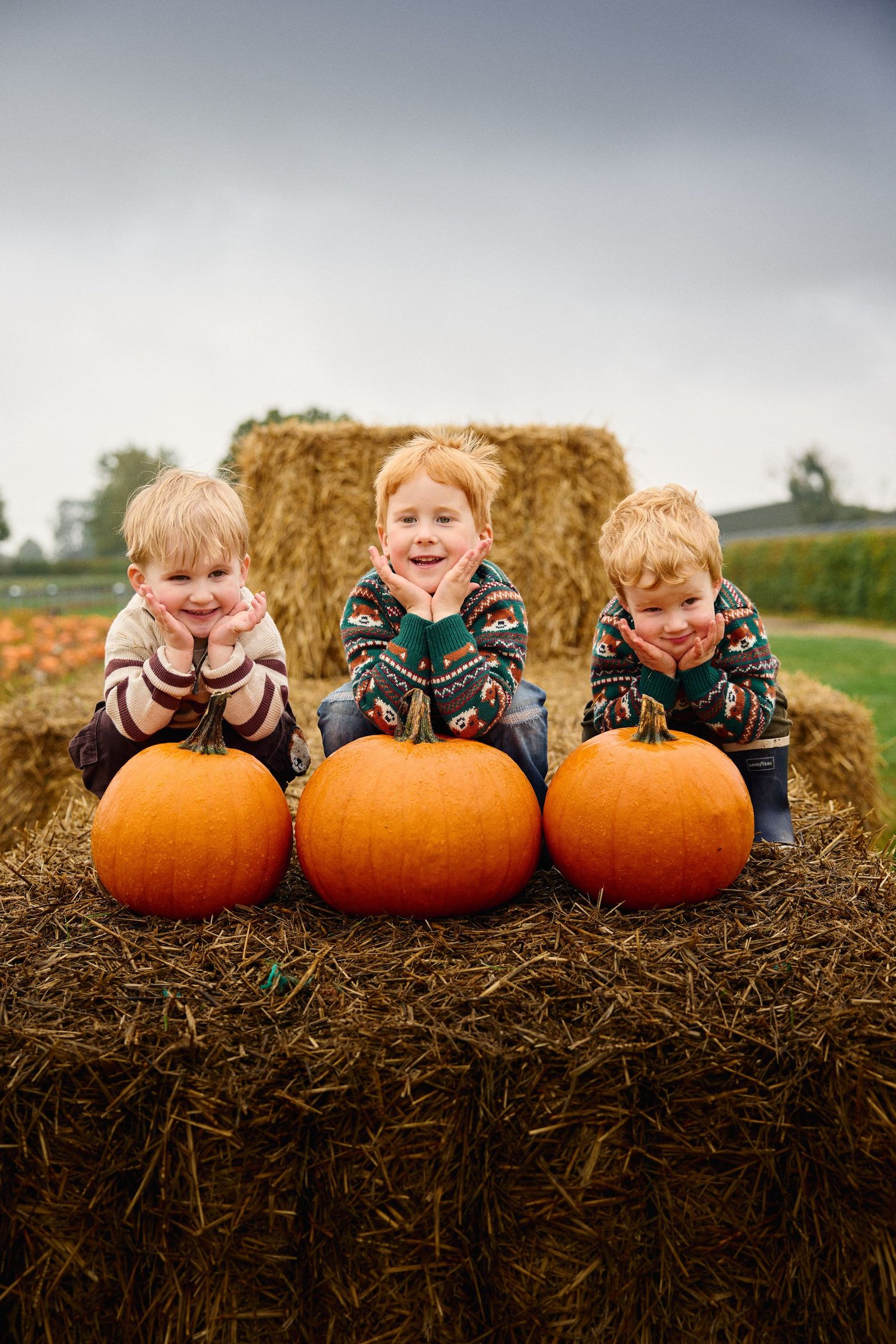 Children lying on hay bales behind three large orange pumpkins, showing excitement.
