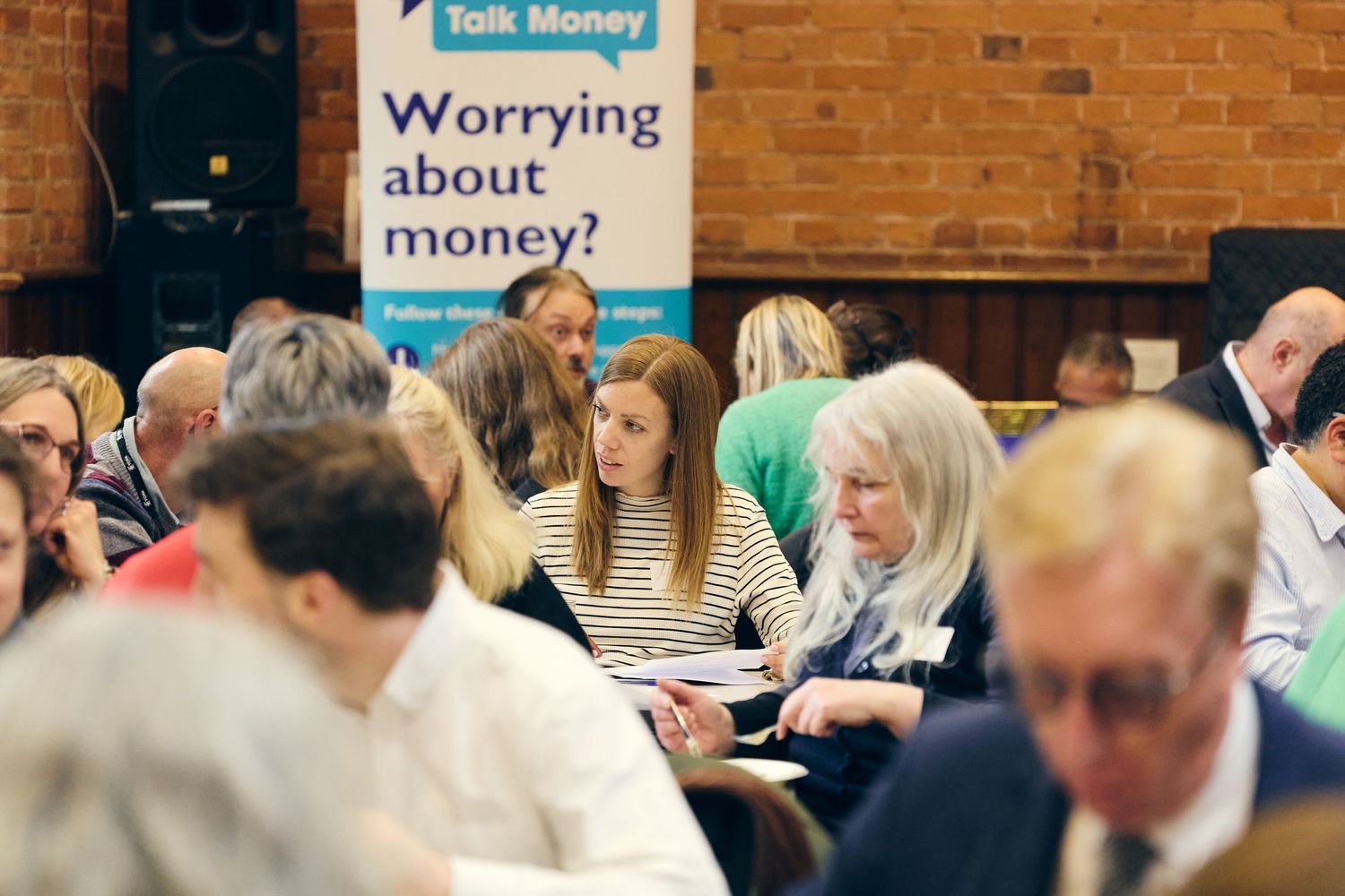 People attending a Talk Money financial wellbeing event, seated at tables discussing money worries in a community hall.