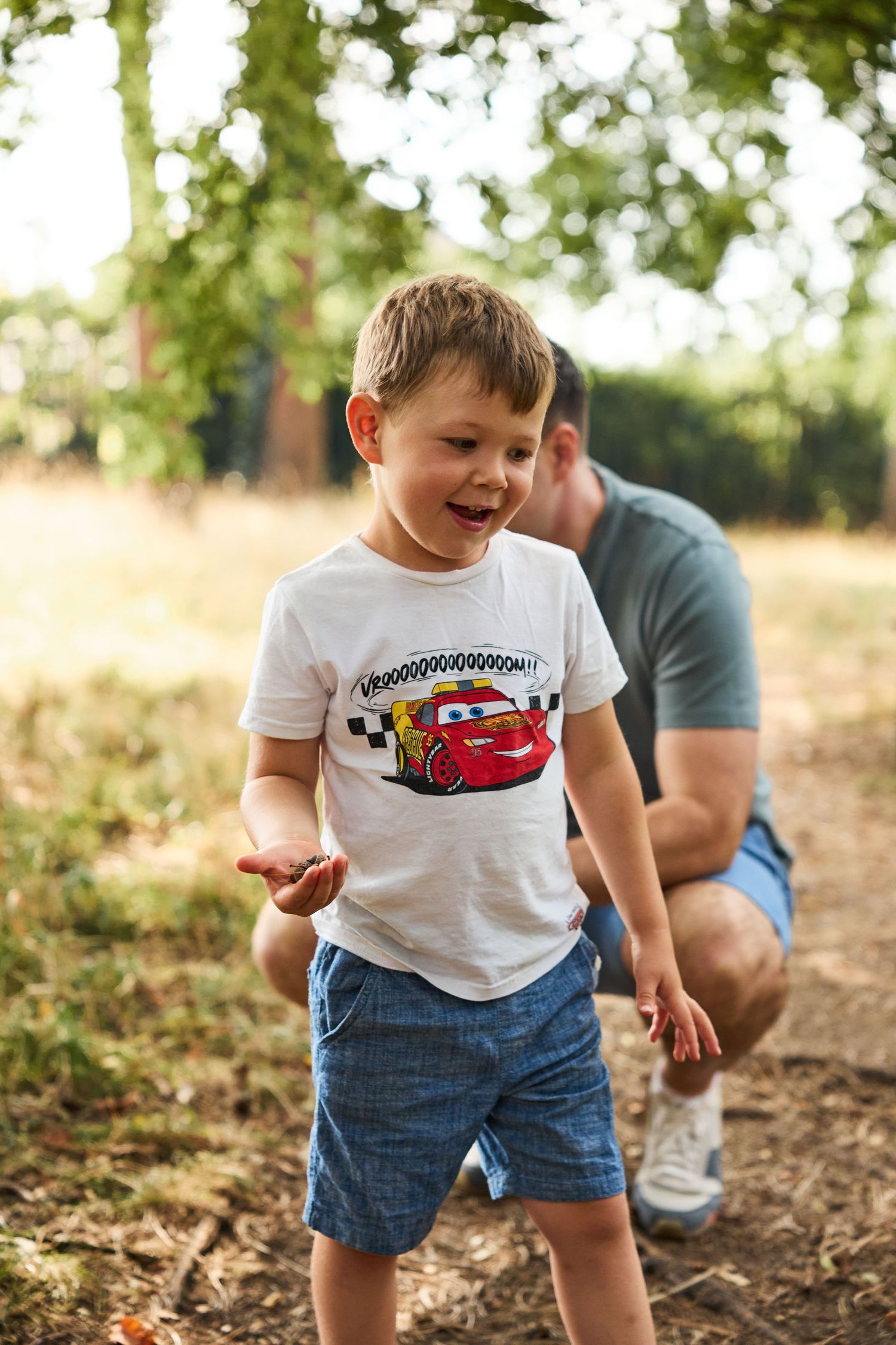 Child wearing a Cars t-shirt plays joyfully on fallen leaves in a wooded setting.