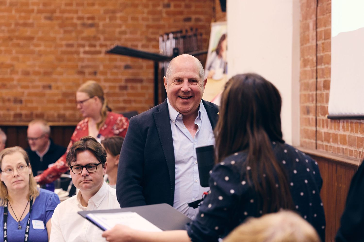 A smiling man in a blue shirt engages with attendees at a professional networking event in a brick-walled venue.
