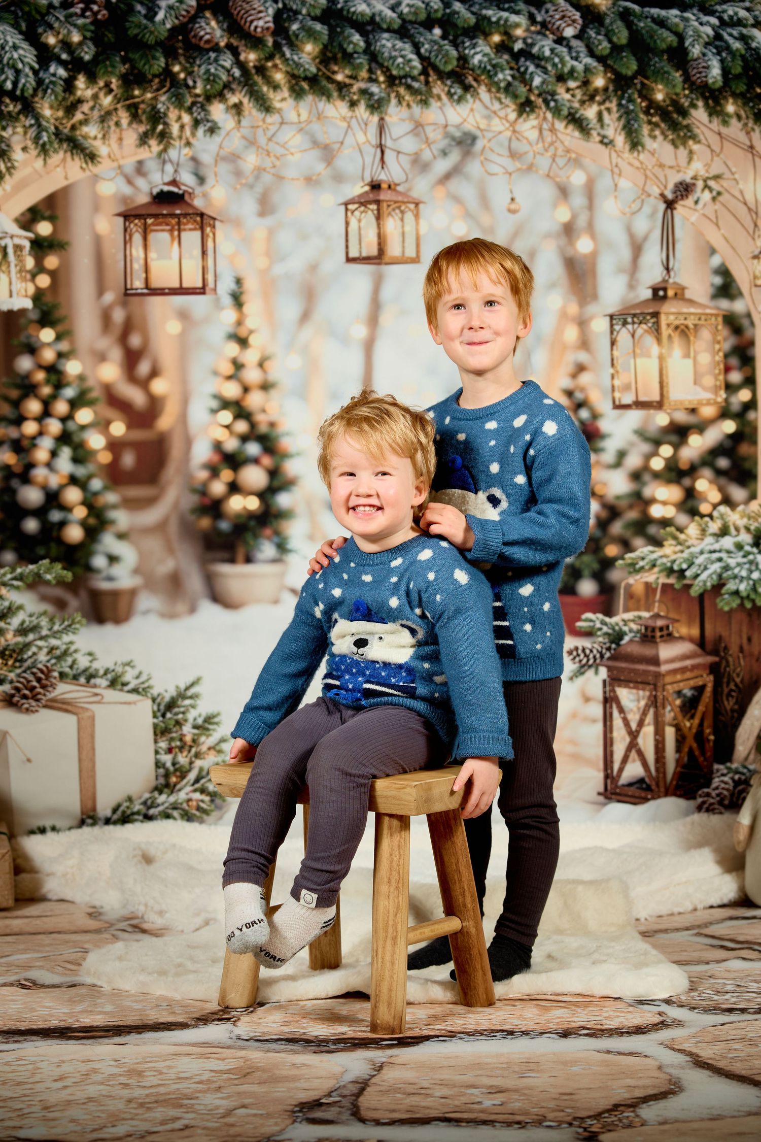 Children in blue sweaters pose on wooden stools against a winter holiday backdrop with lanterns and twinkling lights.