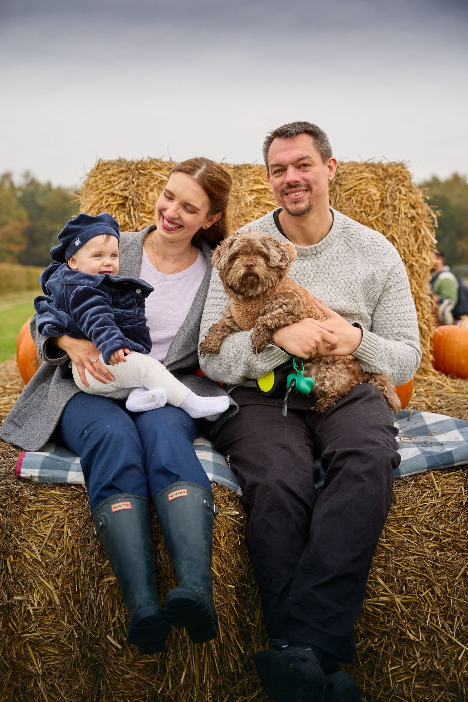 A family and their fluffy dog share a joyful moment while sitting on hay bales at a pumpkin farm.