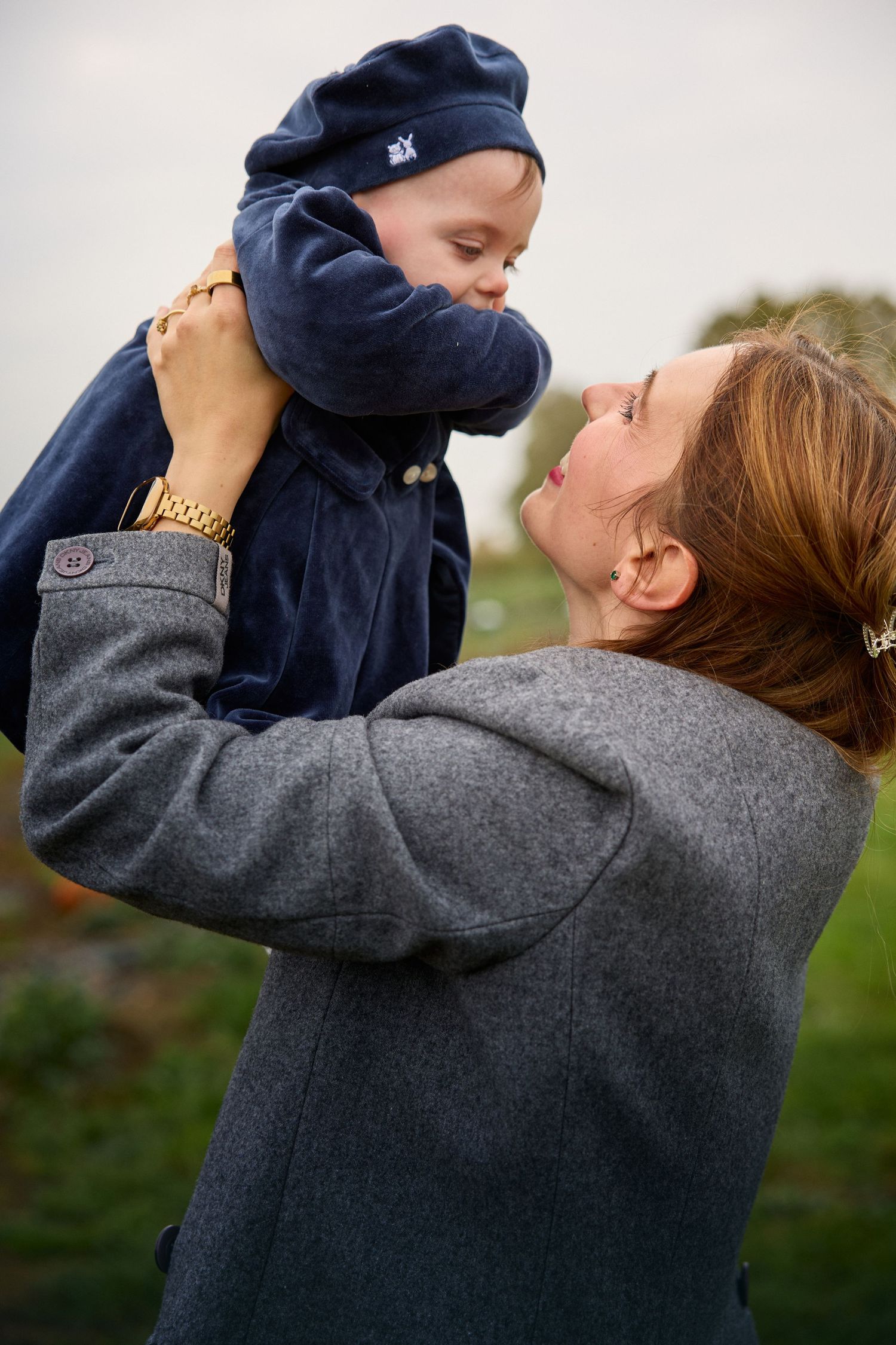 Parent lifts smiling baby in navy hoodie against cloudy sky during outdoor photoshoot.