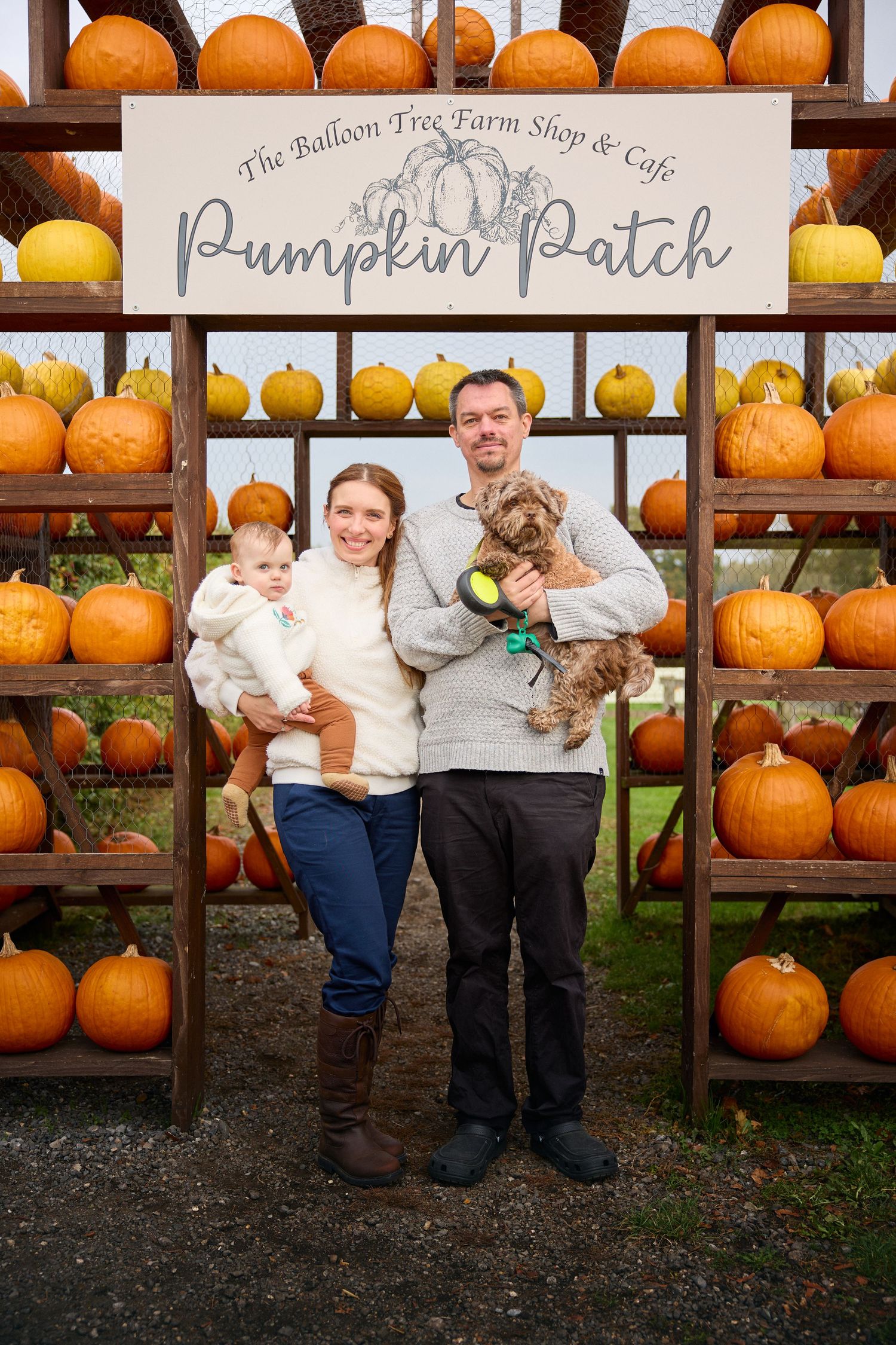 Family poses together in front of wooden shelves filled with bright orange pumpkins at a farm store.