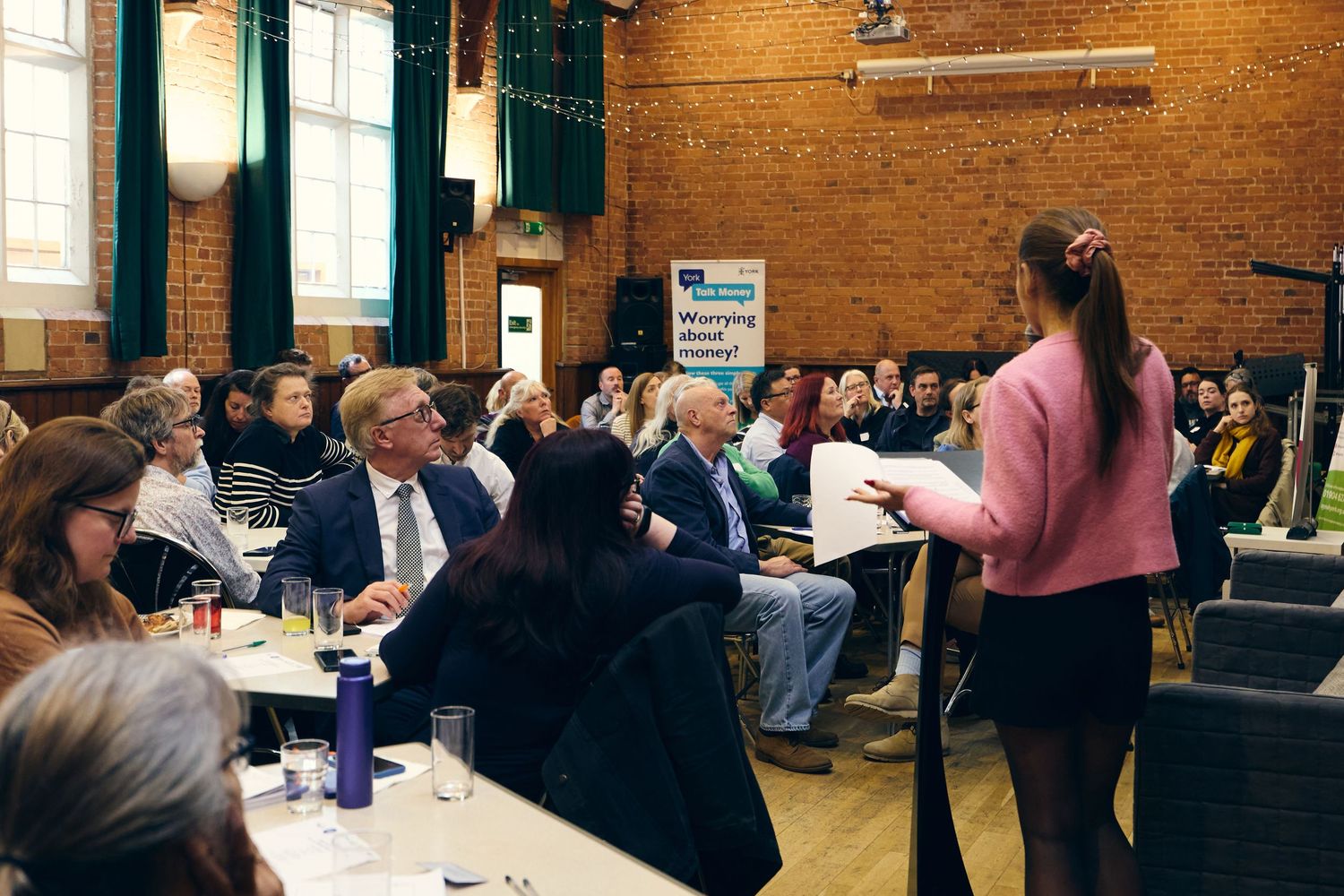 Professional speaker addresses seated audience in brick-walled event hall during a formal conference or community meeting.