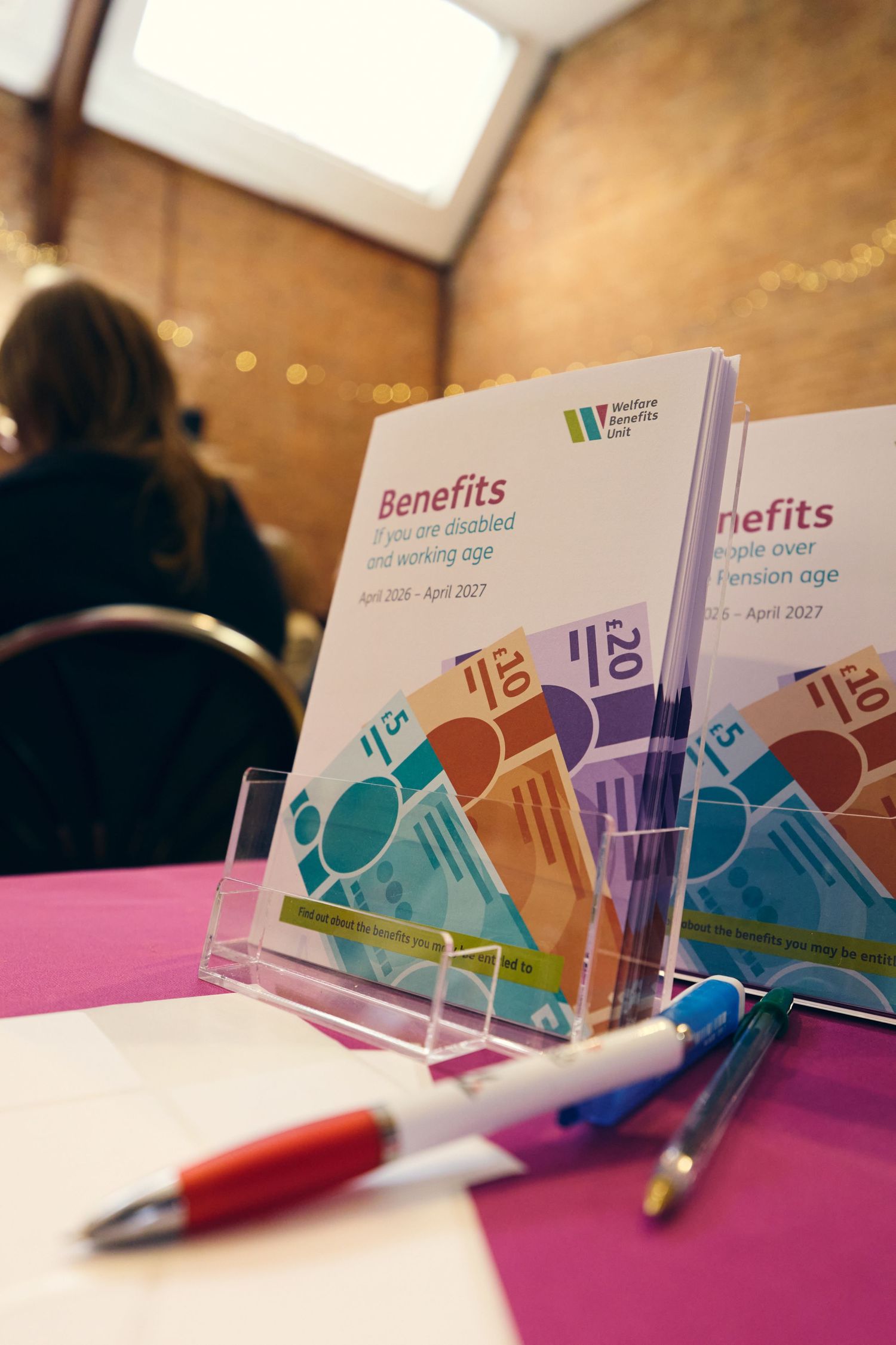 Benefits guides for disabled and pension age people displayed in a clear acrylic stand on a pink table with pens.
