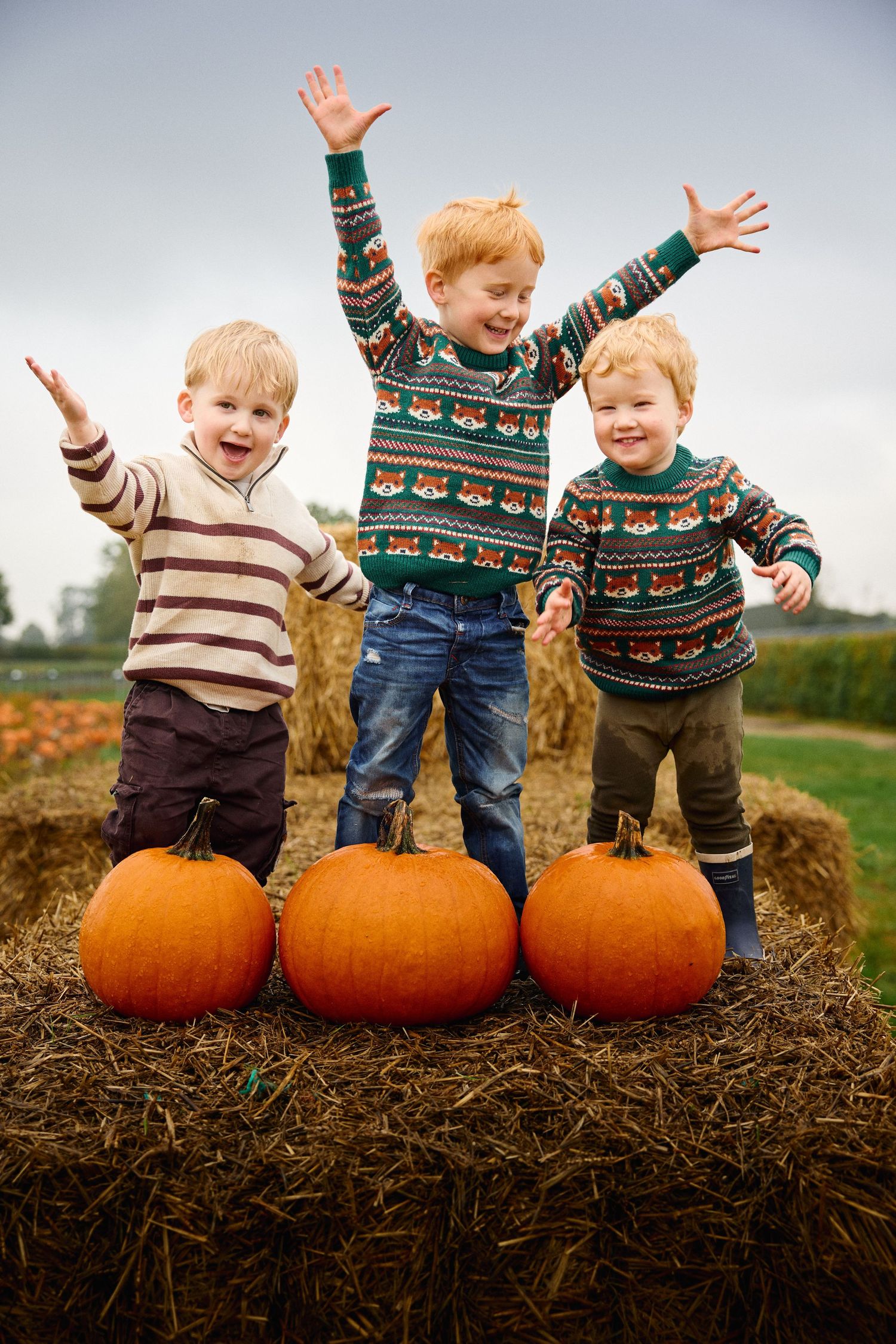 Three children jumping with joy on hay bales behind large pumpkins.