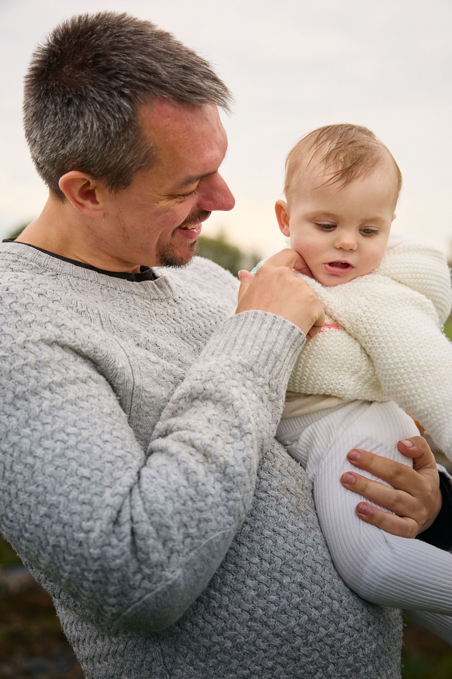 Playful moment between adult in grey sweater and baby in white outfit at pumpkin patch.