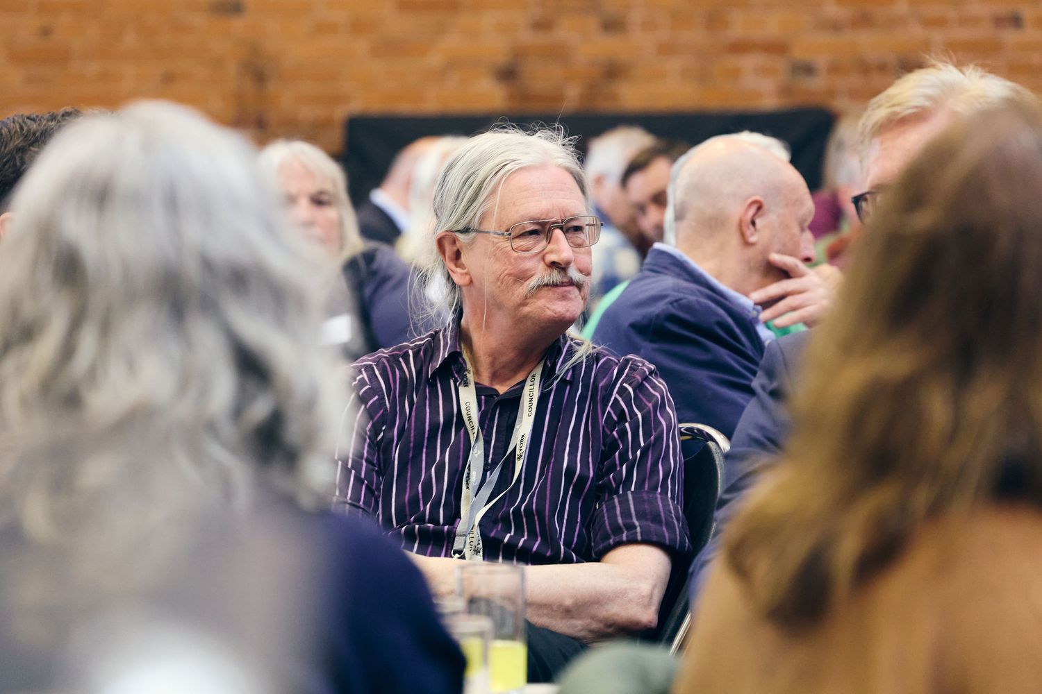 Older man with gray hair and mustache wearing striped shirt and lanyard sits attentively in a crowded conference room.