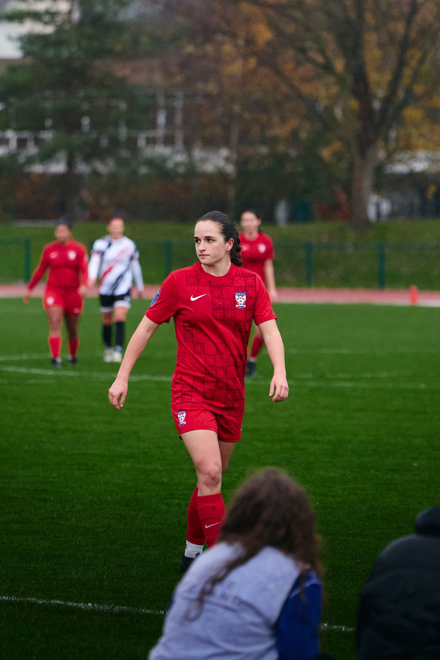 Soccer player in red Nike uniform runs on green field during competitive match with spectators watching from sideline.