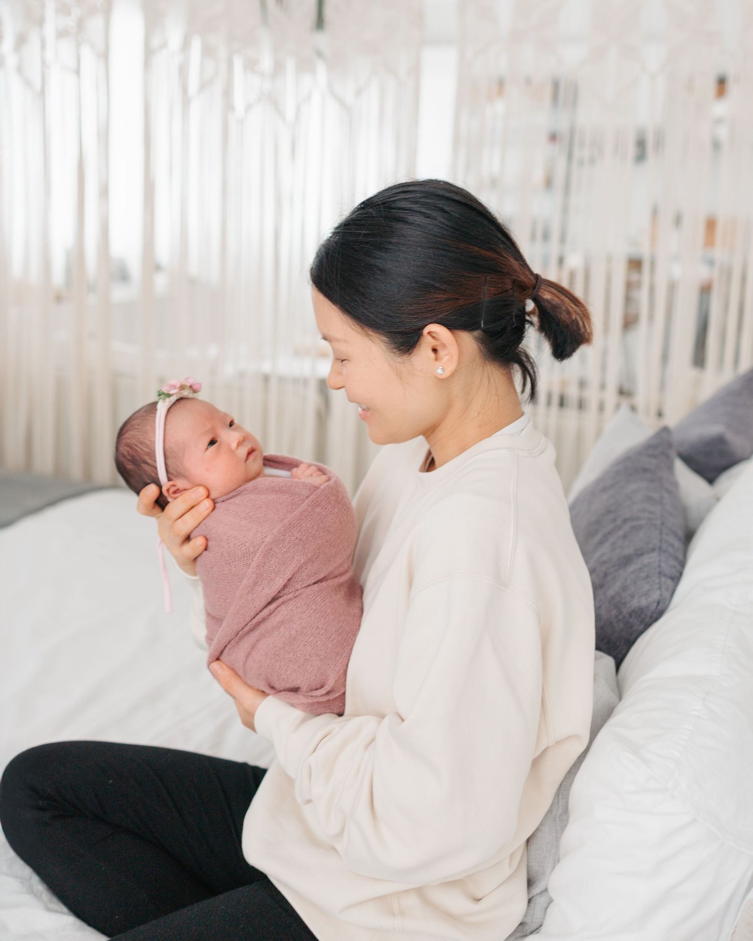 Parent in cream sweater holds and gazes lovingly at newborn baby wrapped in dusty rose swaddle on cozy bed with pillows. Newborn HK