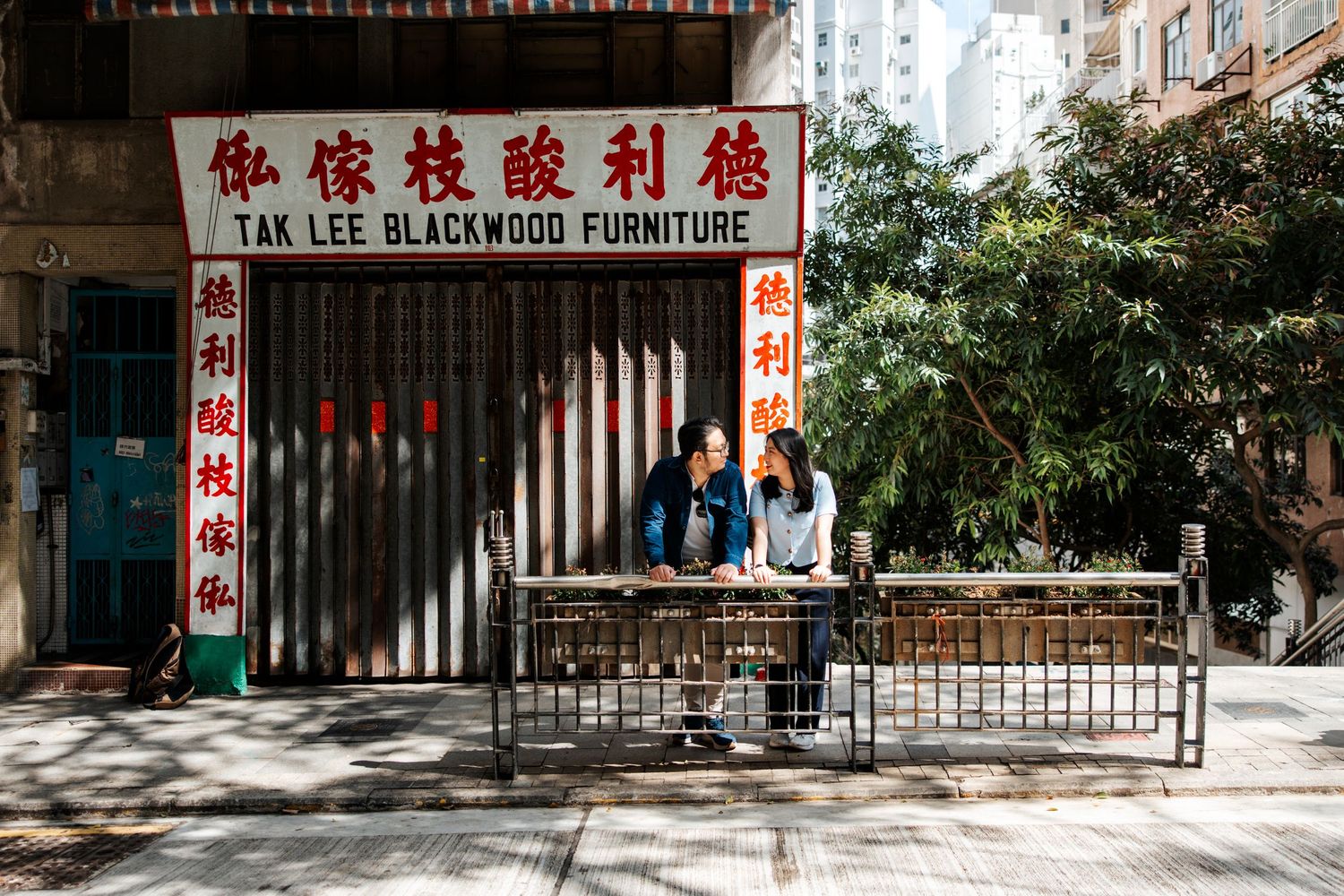 Chinese storefront signs and text adorn a commercial building exterior in an urban setting with trees visible. Couple Photoshoot in HK