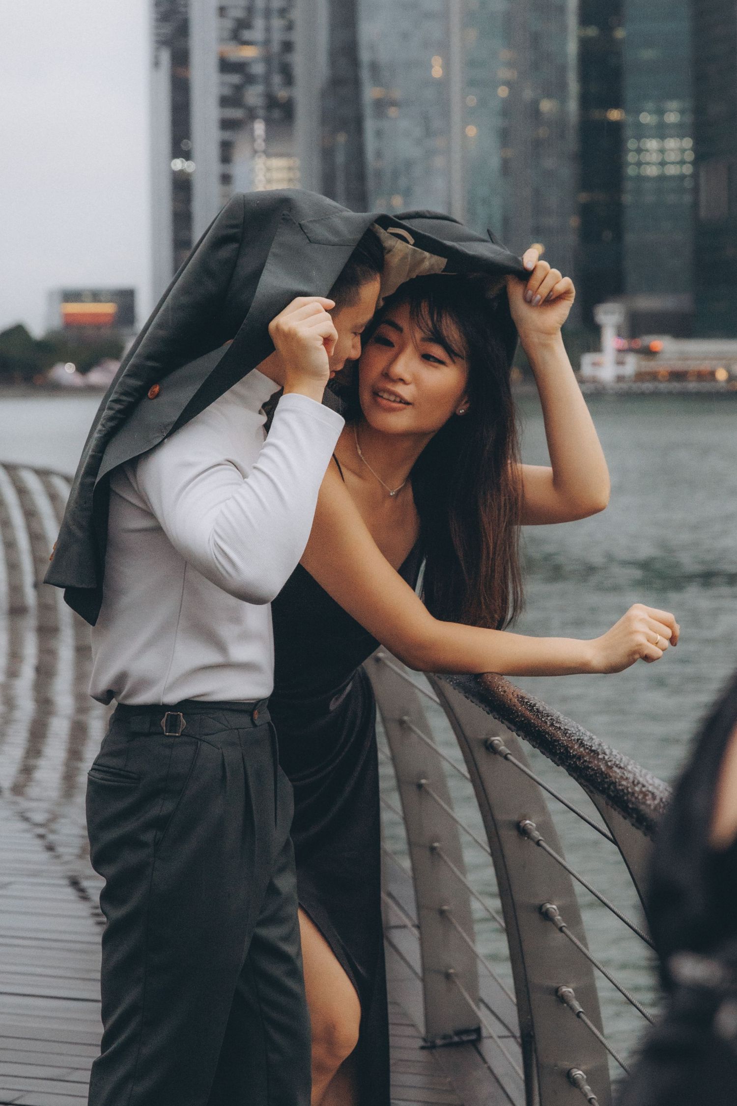 A romantic couple shares an intimate moment under a jacket in the rain and waterfront in the background. Pre Wedding Hong Kong