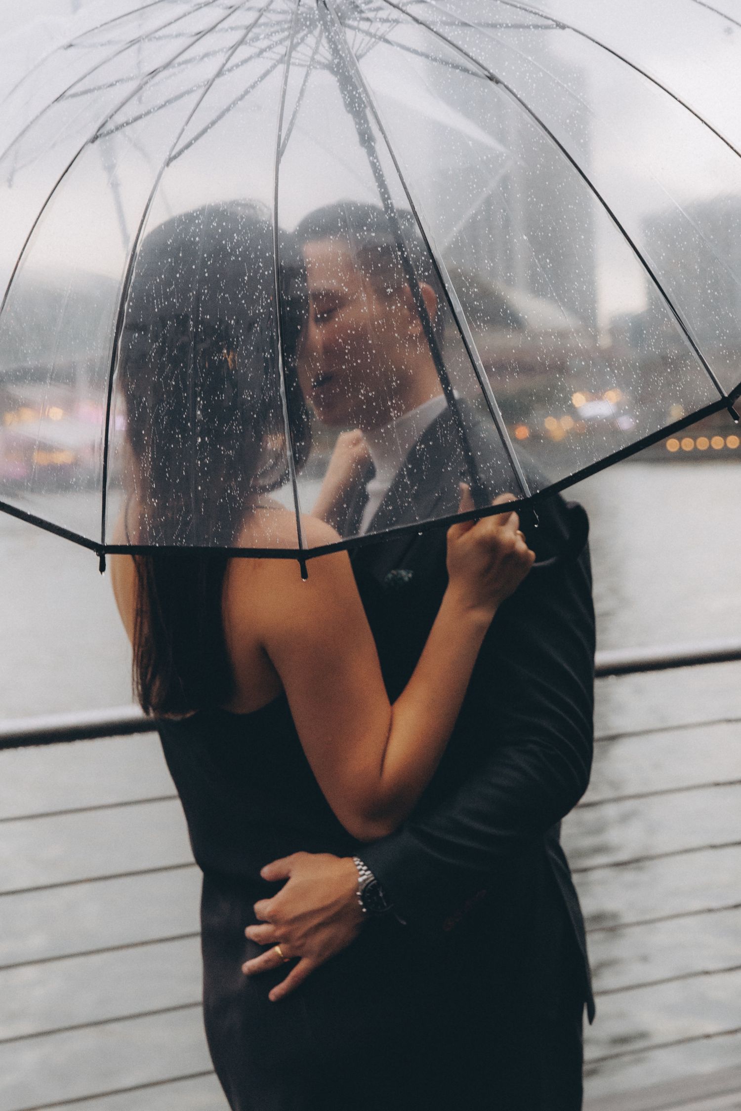 A romantic couple shares an intimate moment under a clear umbrella in the rain with city lights in the background. Couple Photoshoot in HK