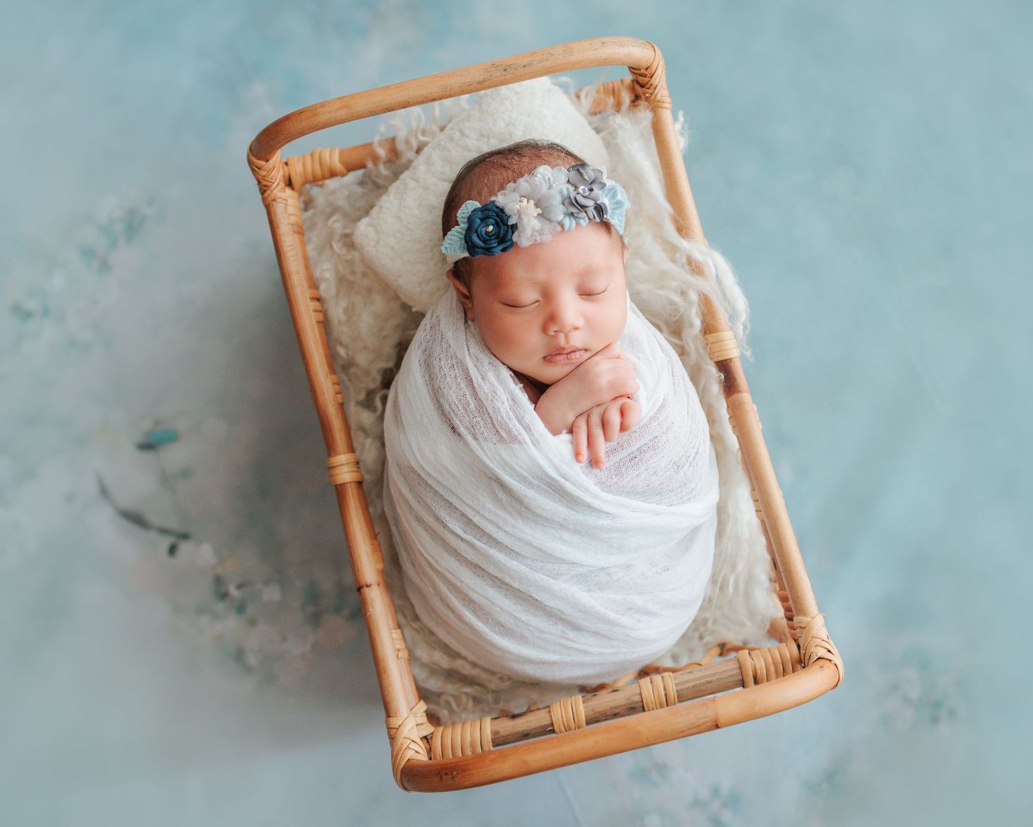 Baby wrapped in white swaddle blanket sleeps peacefully in wicker basket on turquoise backdrop. In-home newborn session in Hong Kong