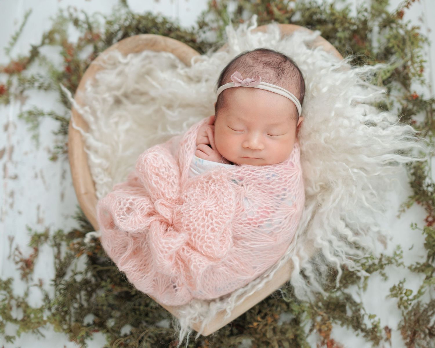 A newborn swaddled in delicate pink wraps sleeps peacefully on soft white fur and woodland accents. Newborn Photographer in Hong Kong