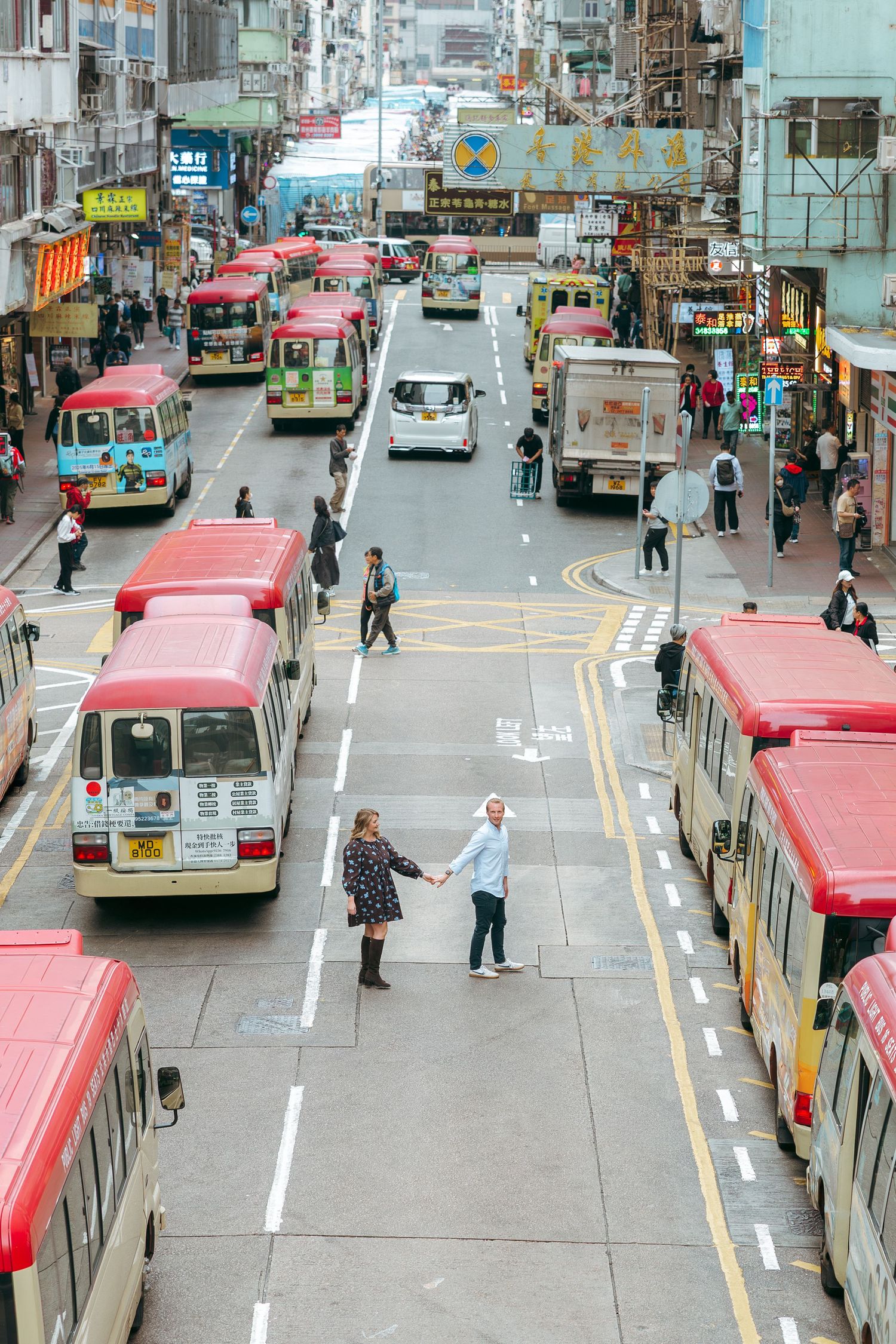 Couple holds hands on a busy Hong Kong street surrounded by red-roofed buses and city traffic. couple session in Hong Kong. Pre Wedding HK