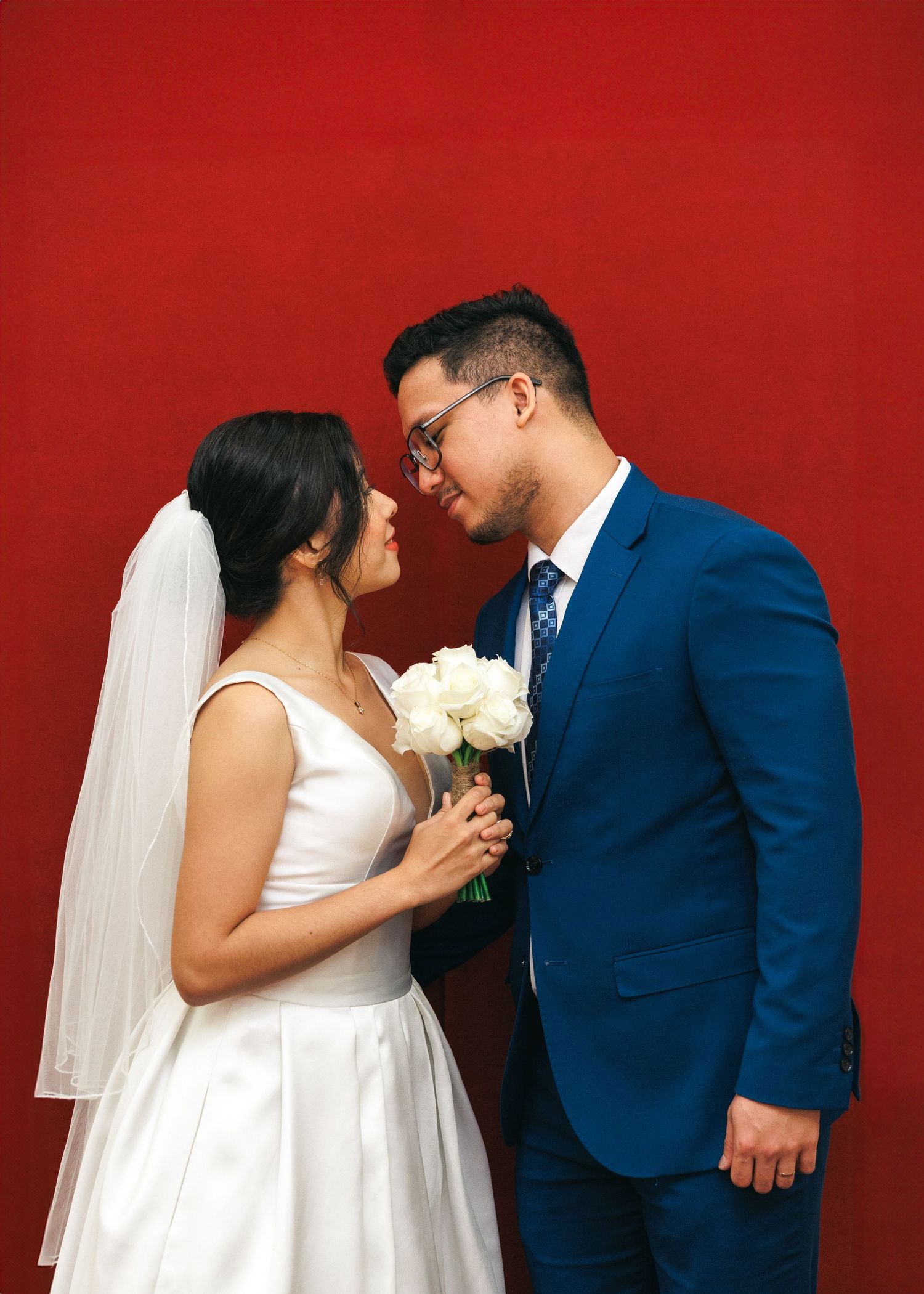 A couple on their wedding day sharing an intimate moment against a bright red backdrop, with the bride holding white roses. Civil Ceremony