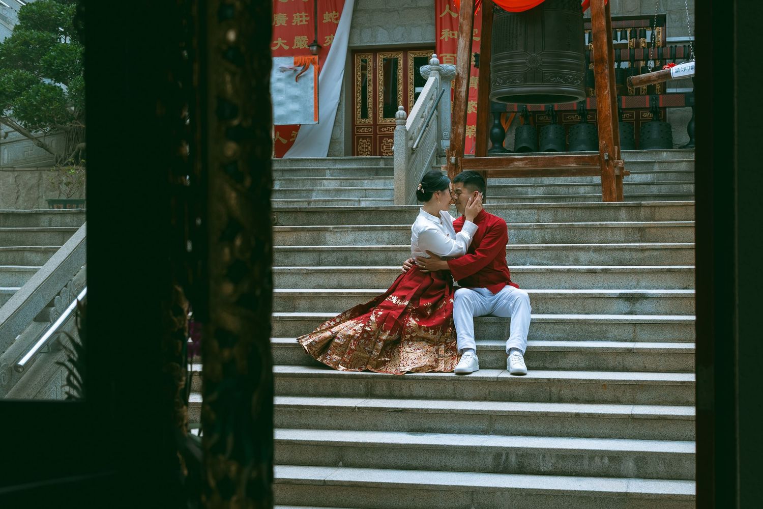 couple n traditional Asian wedding attire sit together on stone steps adorned with red lanterns and decor. Couple Photoshoot in Hong Kong