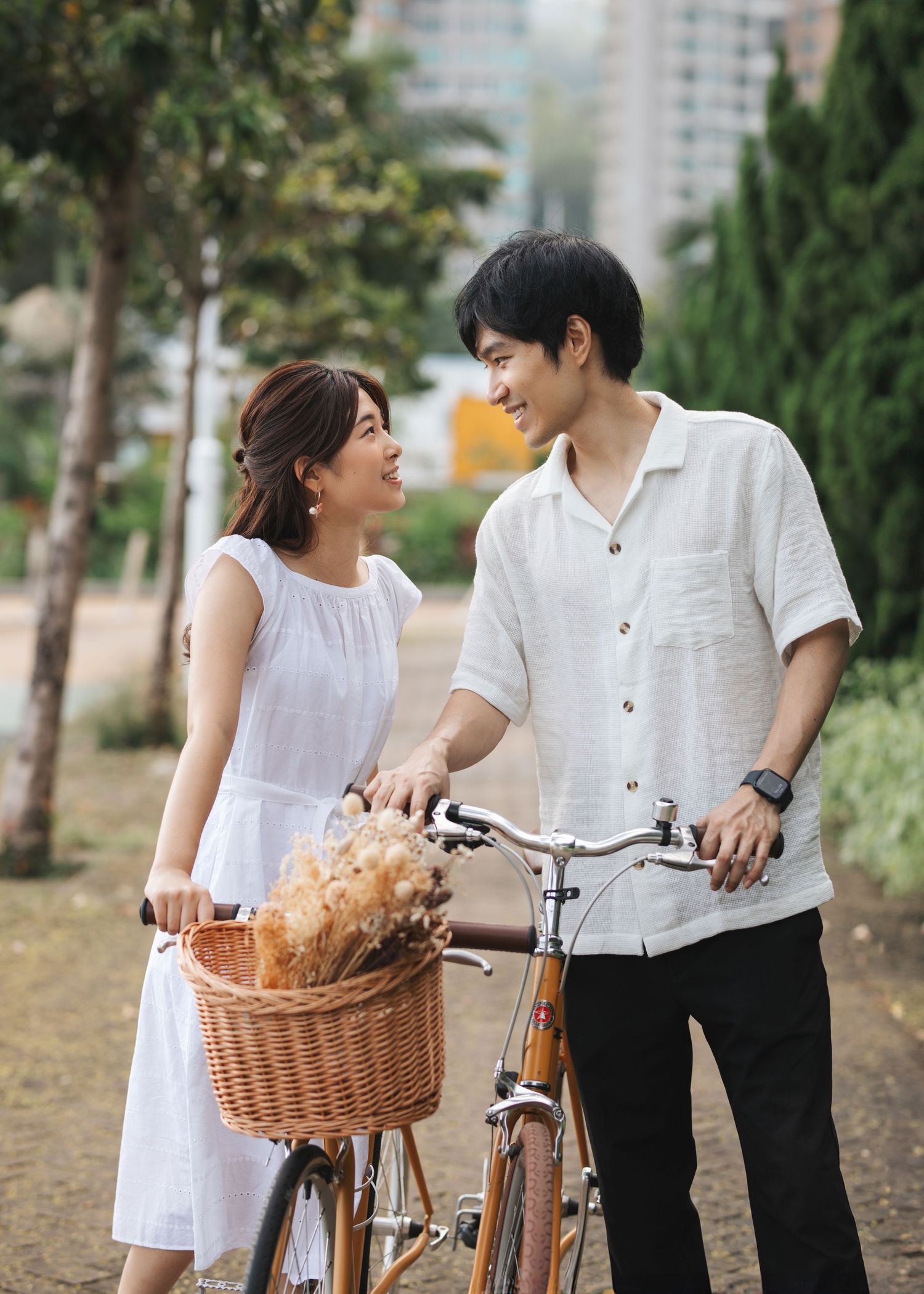 A couple in white and black outfits walking with a bicycle and wicker basket in an outdoor park setting. Couple Photoshoot in Hong Kong