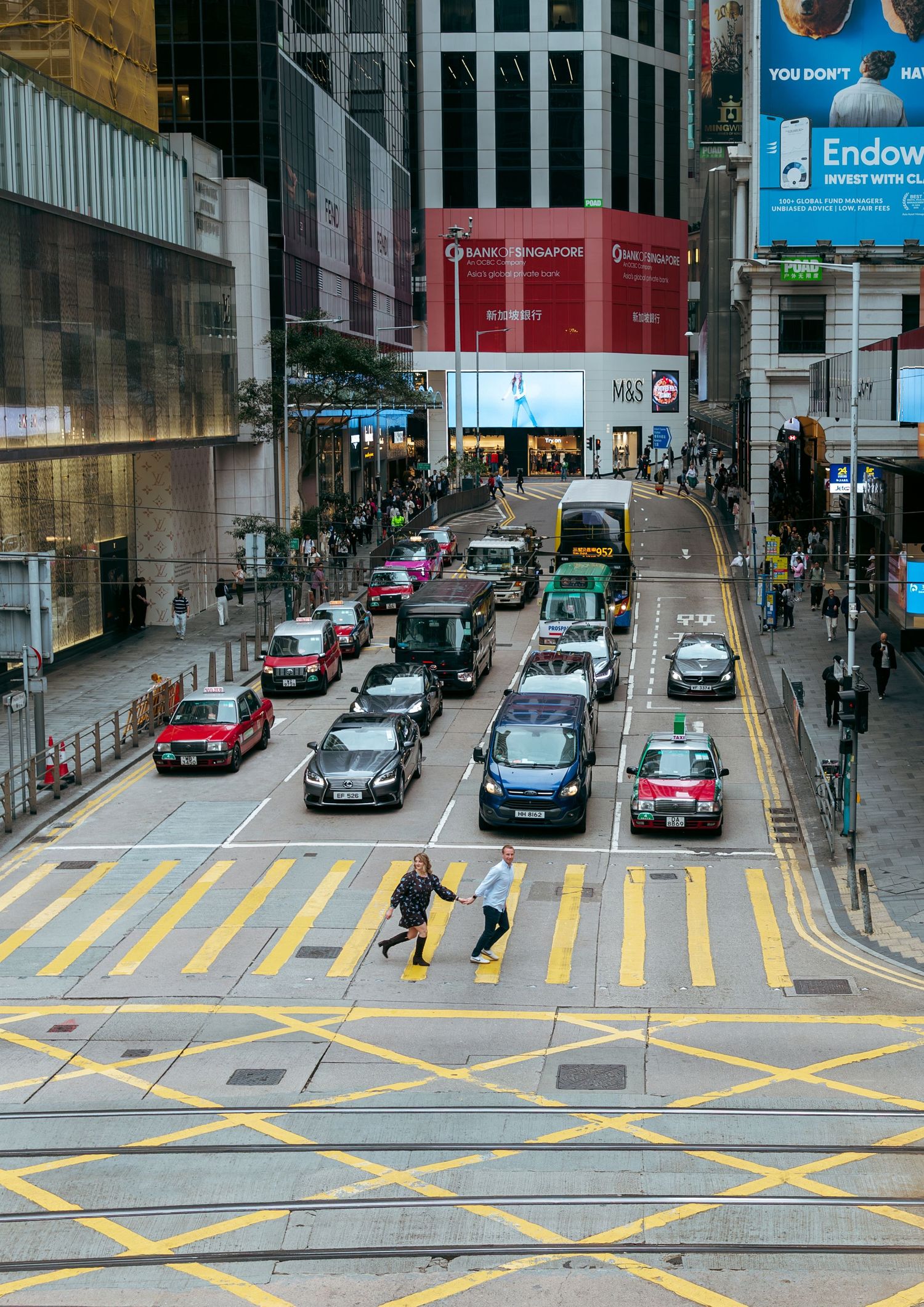 Busy Hong Kong street intersection with yellow crosswalk, taxis, and pedestrians crossing amid tall commercial buildings. couple session