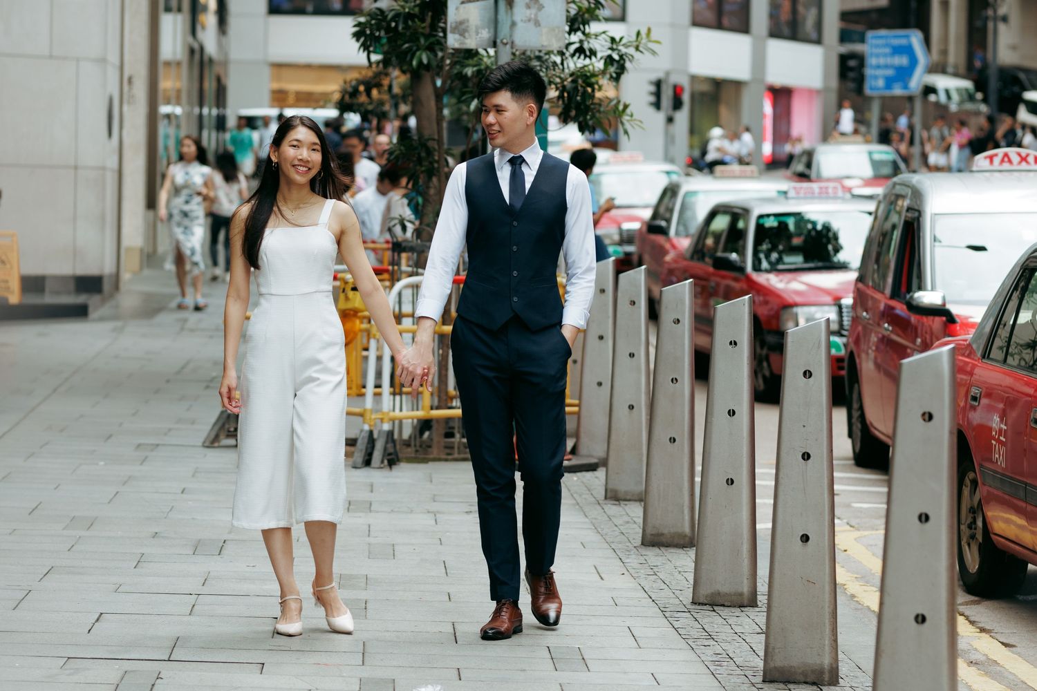 A couple in formal wedding attire walks on a city sidewalk holding hands among parked cars and pedestrians. Couple Photoshoot in Hong Kong