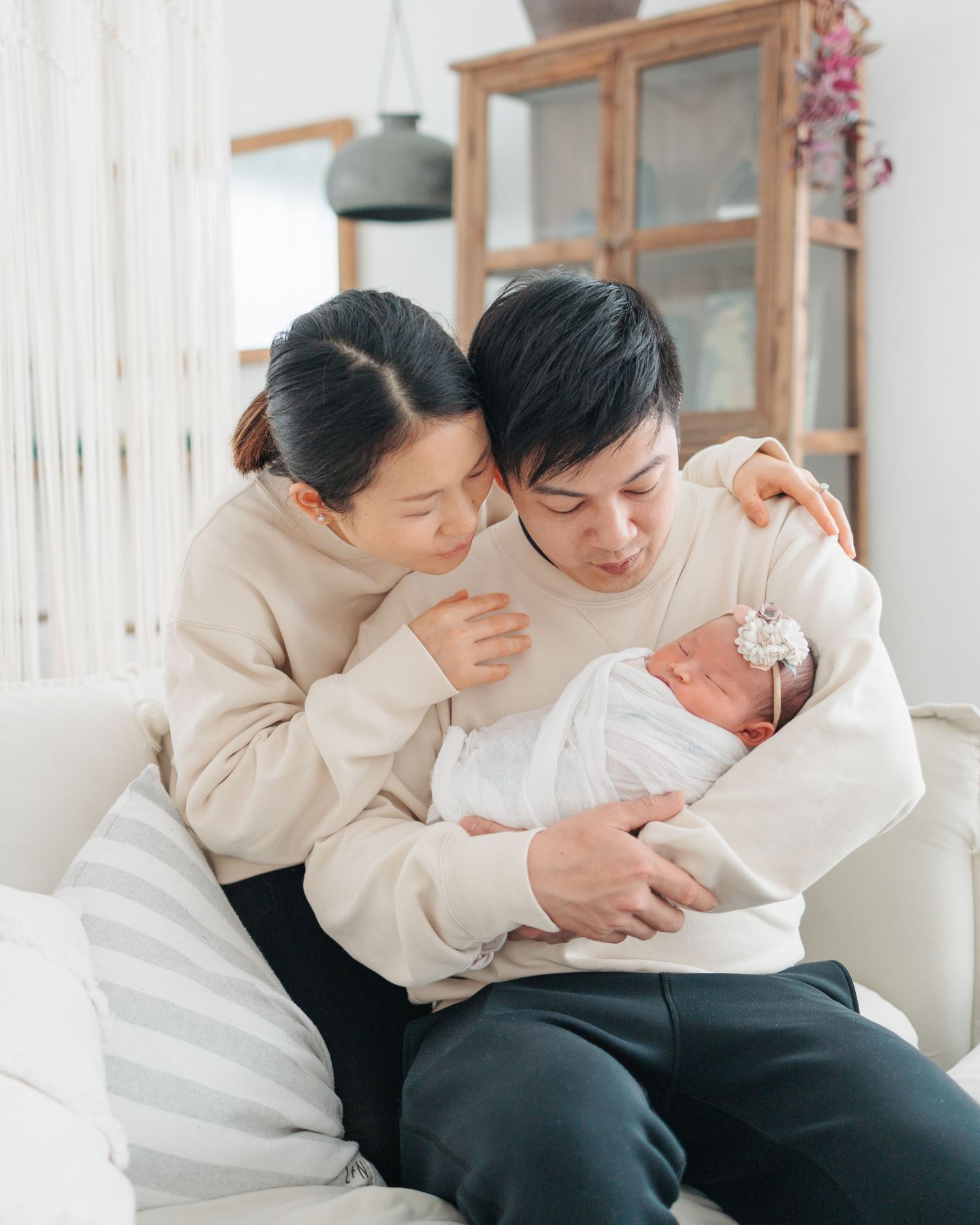 Couple in matching beige sweaters gaze lovingly at their newborn baby wrapped in white blanket on sofa. Newborn Photographer in Hong Kong