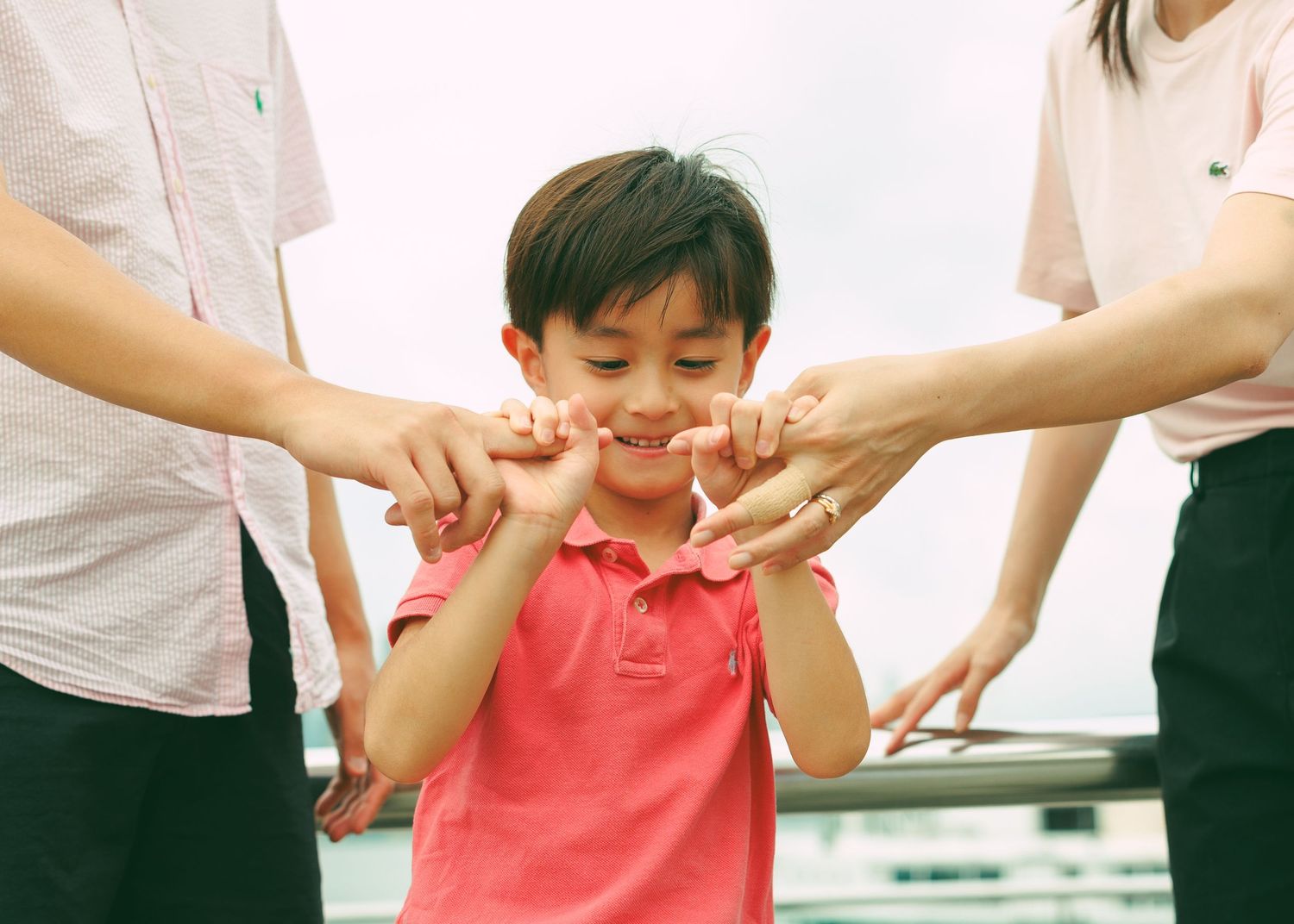 Someone wearing a coral pink t-shirt smiles while adults playfully squeeze their cheeks in a candid moment. Family Photoshoot in Hong Kong