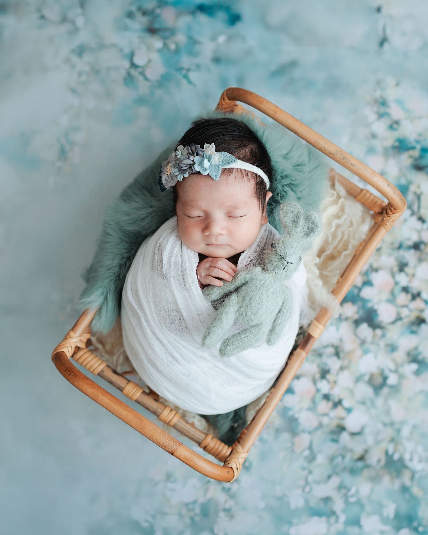 A sleeping newborn wrapped in white fabric rests on a small wooden bed with soft teal floral backdrop. Newborn Photoshoot in Hong Kong
