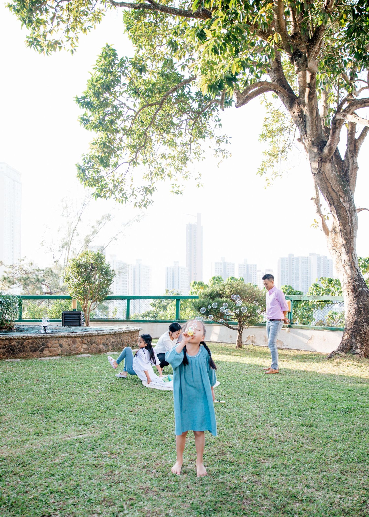 People enjoy a relaxing afternoon on grassy hillside overlooking Hong Kong's iconic skyline through tree branches. Hong Kong Family Photogra