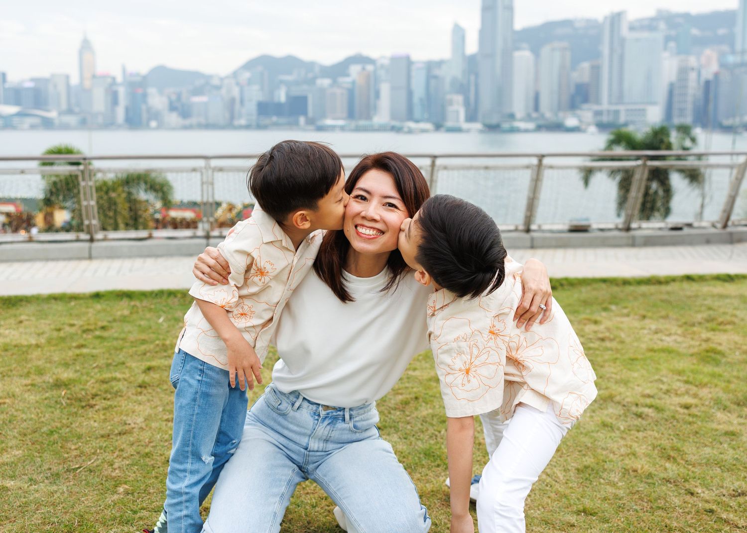 A family sharing a joyful moment together on a grassy area with Hong Kong's iconic skyline visible across the harbor. Hong Kong Family Photo