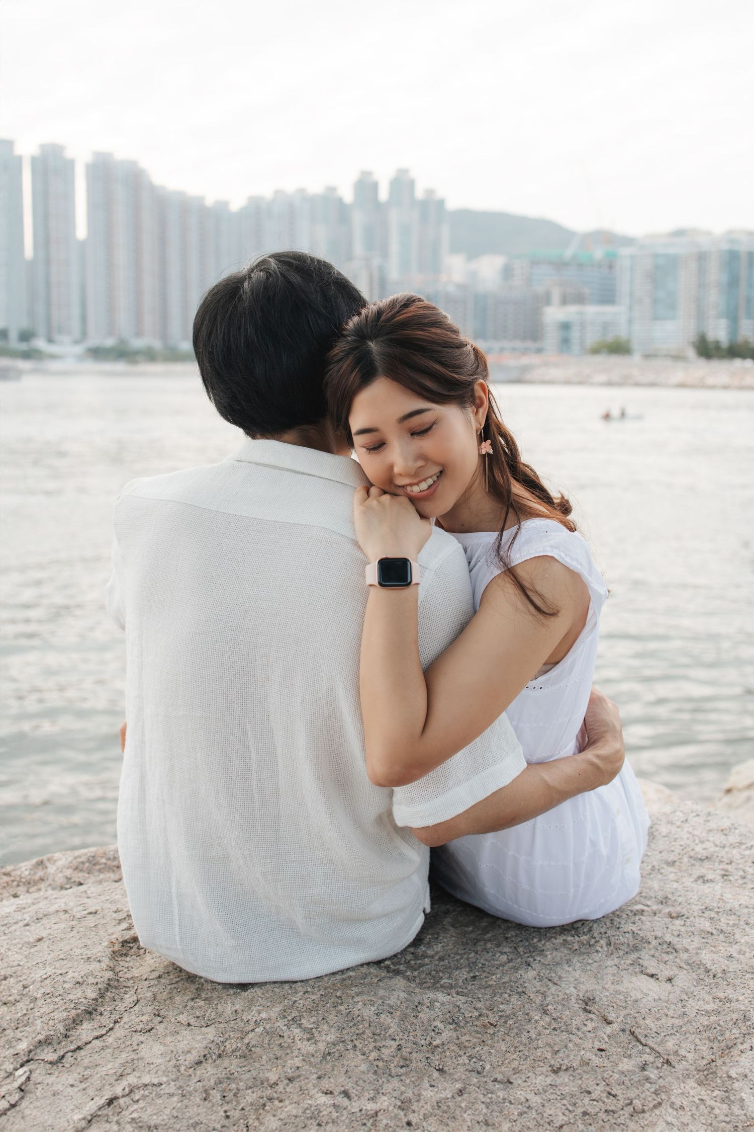 A couple sits closely together on a beach with city buildings visible across the water in the background. Couple Photoshoot in Hong Kong