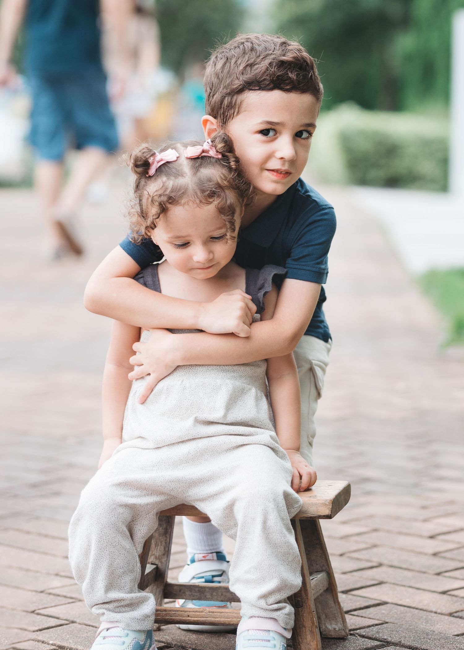 Two children share a tender moment sitting on a wooden bench outdoors, with the older one hugging the younger from behind. Family Photograph