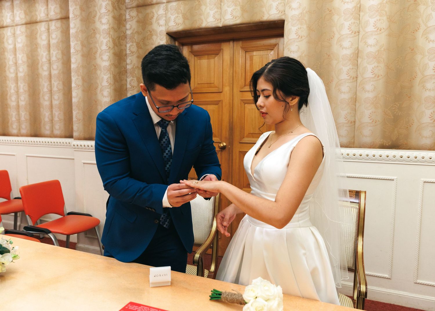 A couple in formal wedding attire sits at a table and signs documents during their ceremony. Civil Ceremony in Hong Kong