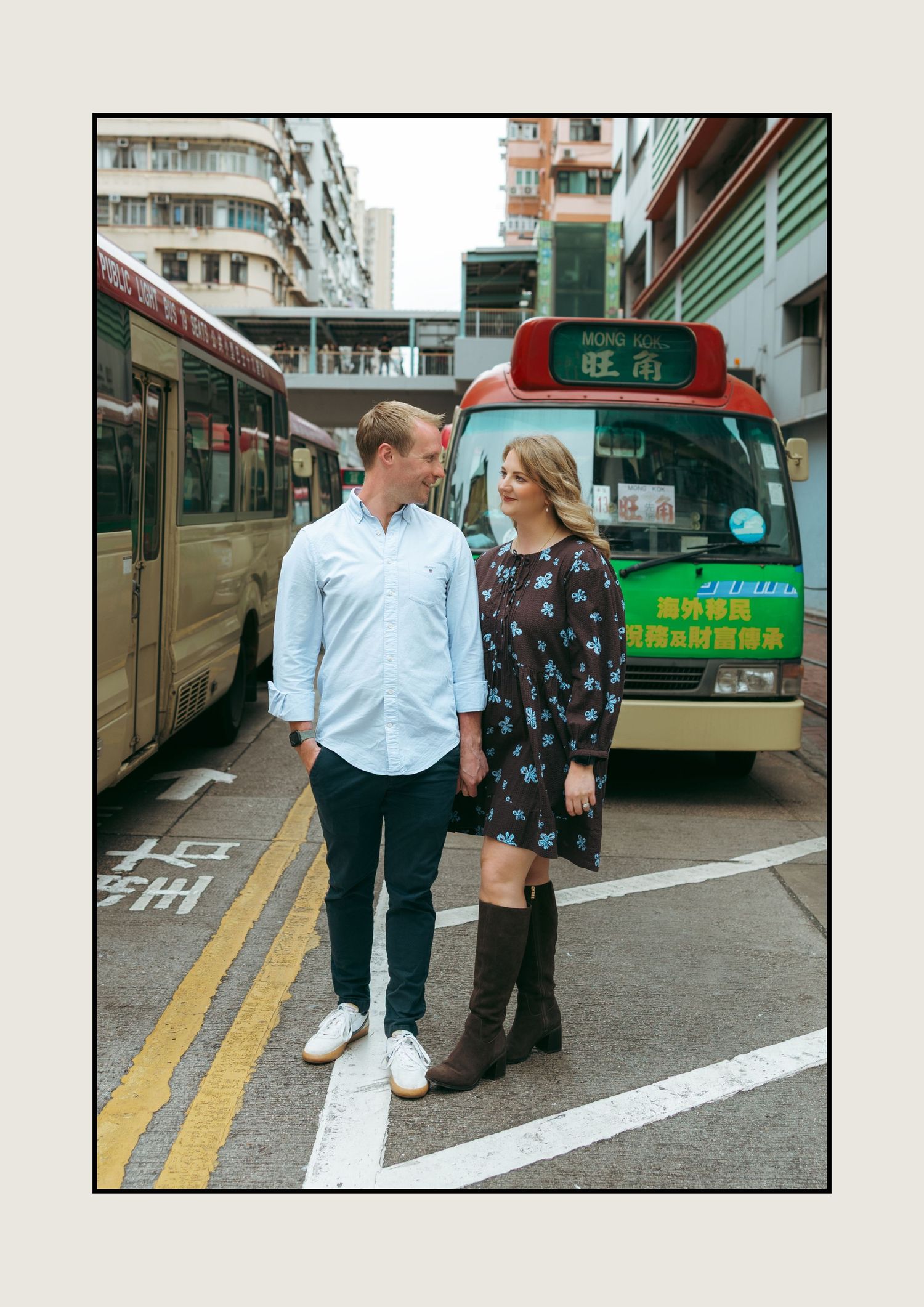 Couple standing on a street crosswalk in Hong Kong with colorful buses and Chinese signage in the background. couple session in Hong Kong.