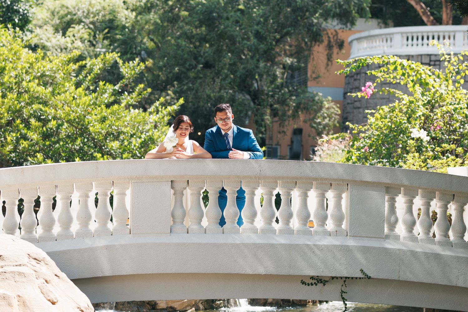 A couple stands together on an elegant white stone balustrade bridge in a lush garden setting on a sunny day. Civil Ceremony in Hong Kong