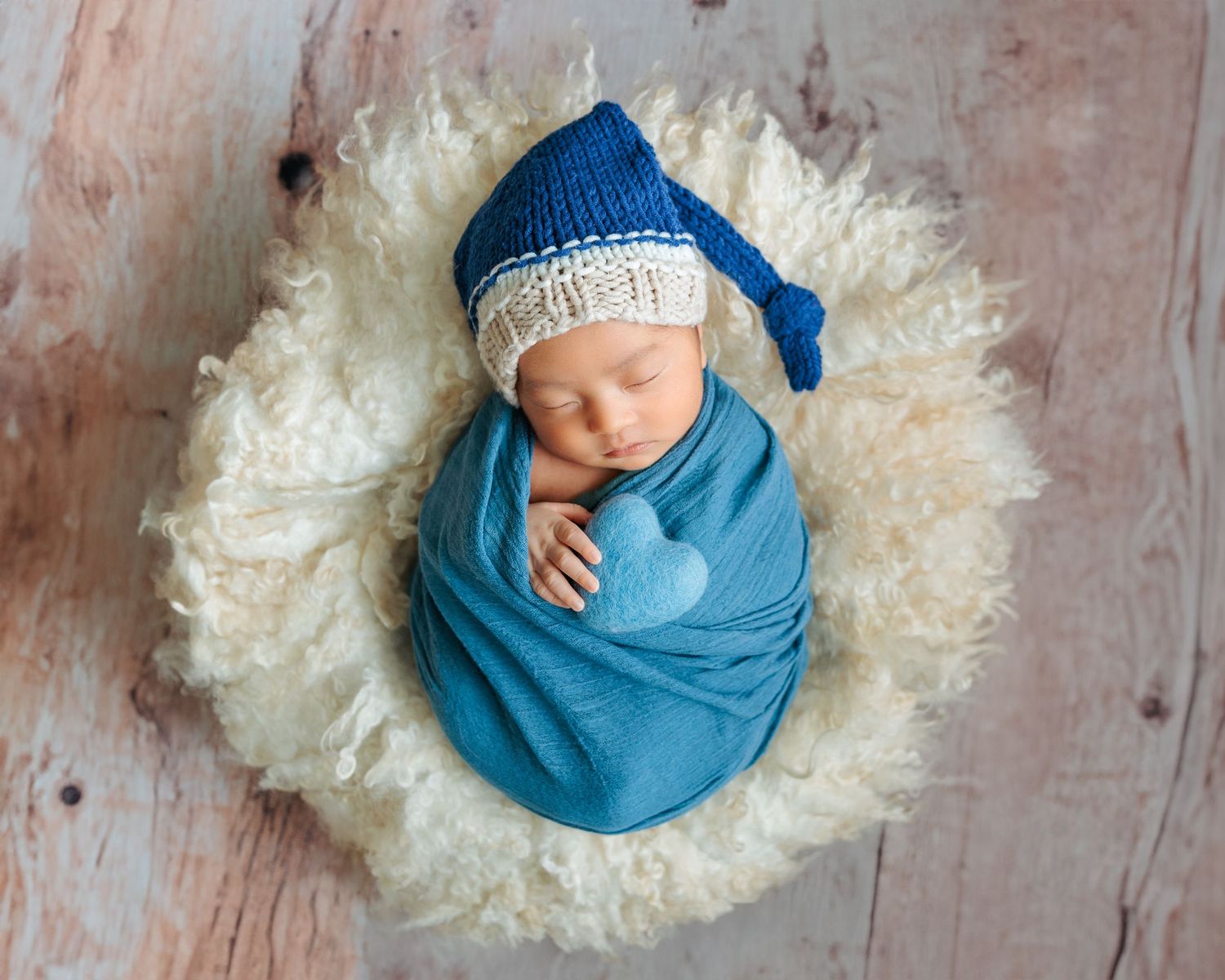 A sleeping newborn wrapped in soft blue fabric lies on a cream-colored furry blanket in a minimalist photo setup. Newborn Photoshoot