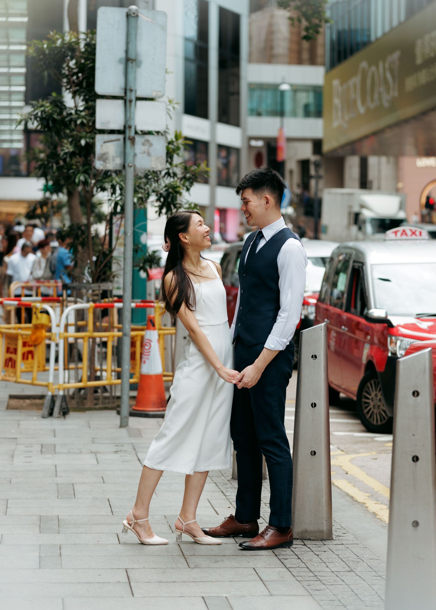 couple in formal attire share an intimate moment on a busy city sidewalk with metal bollards in the foreground. Couple Photoshoot in HK