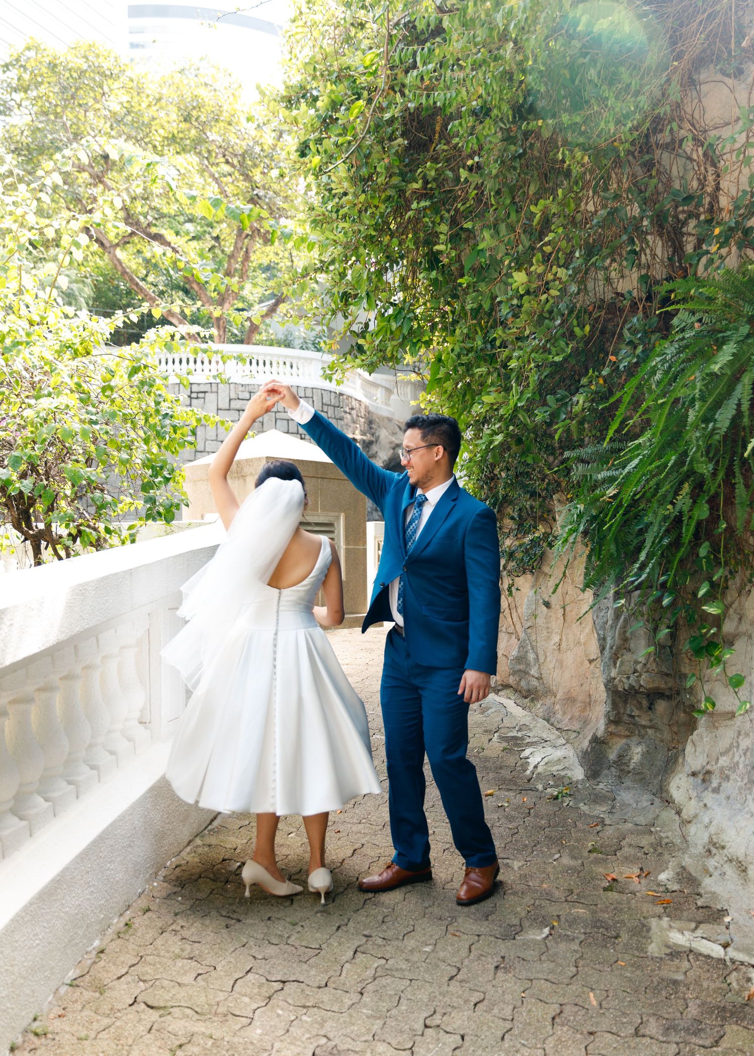 A couple in formal attire playfully dances and twirls together on a stone pathway surrounded by lush greenery. Civil Ceremony in Hong Kong