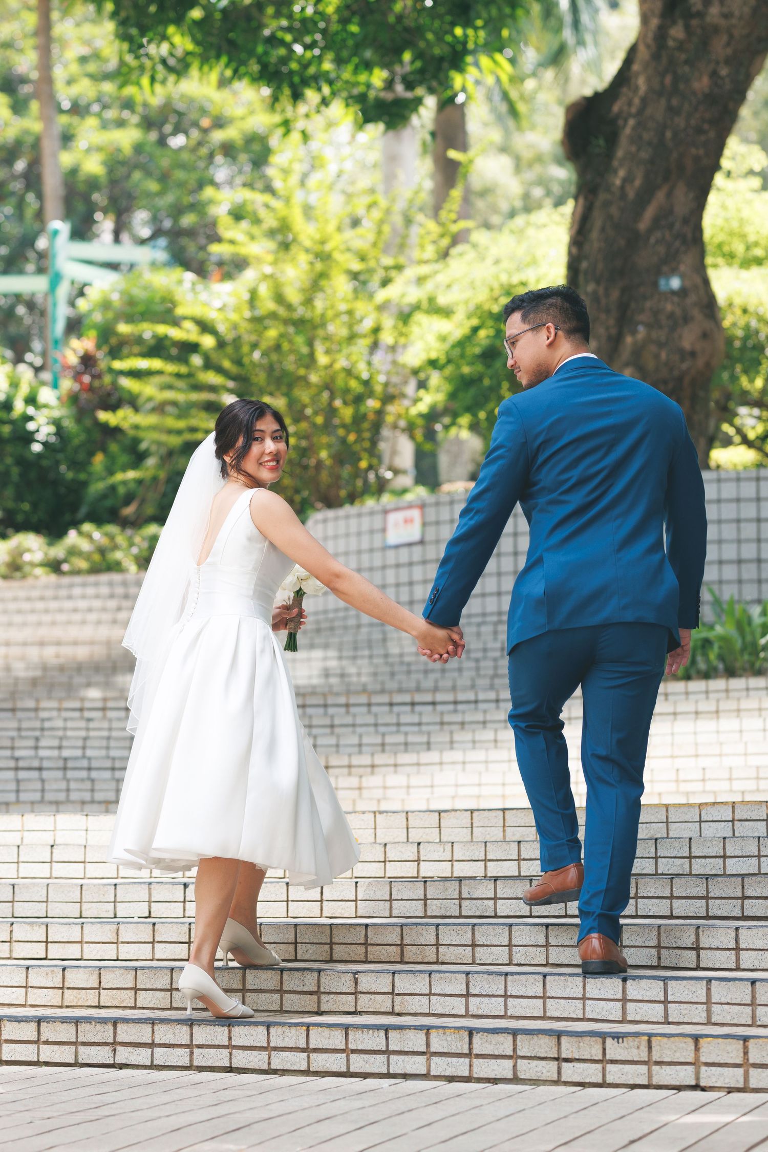A couple in wedding attire holding hands while walking up stairs in a lush outdoor park setting. Civil Ceremony in Hong Kong