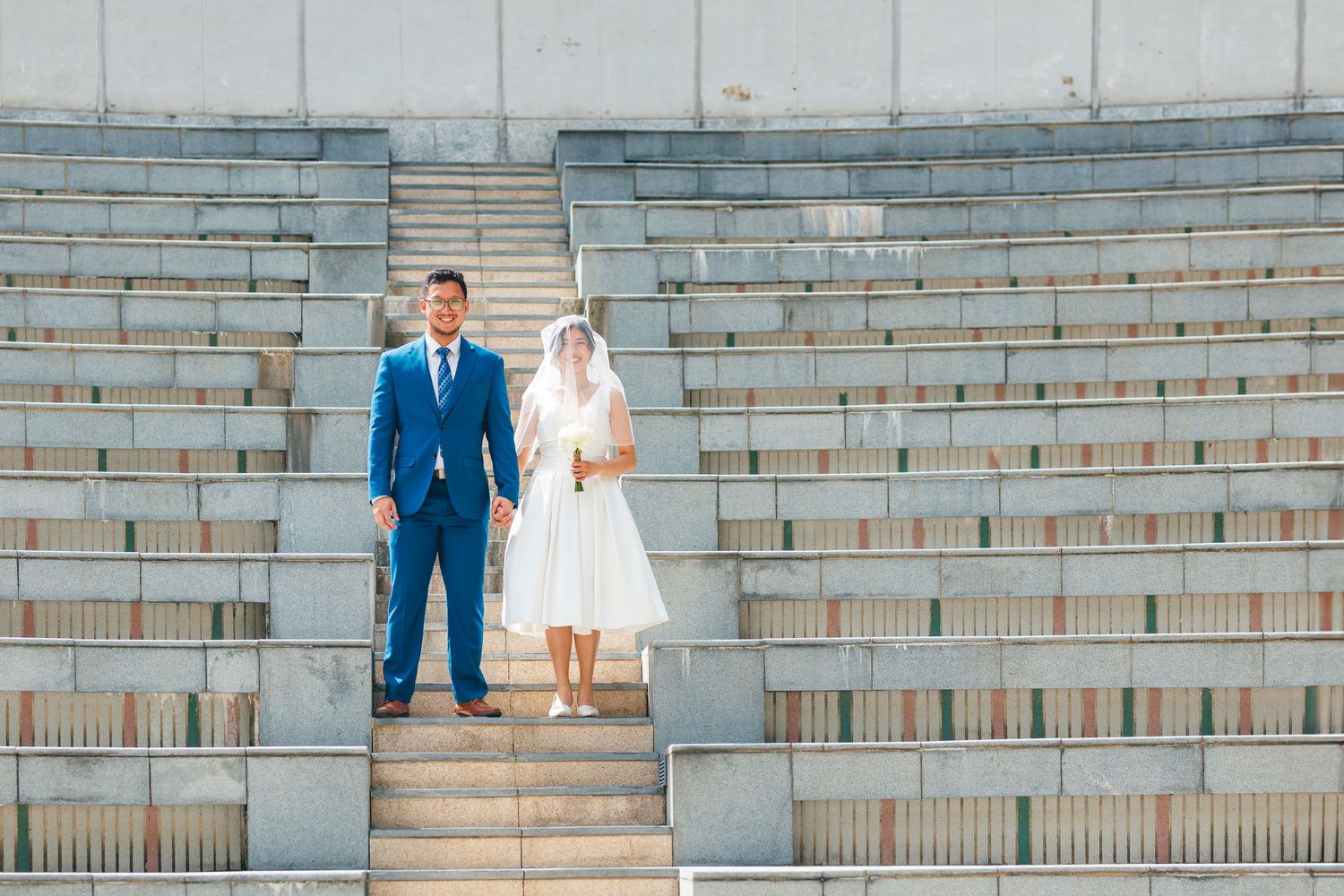 A couple dressed in blue suit and white dress stand together on concrete stadium steps. Civil Ceremony in Hong Kong