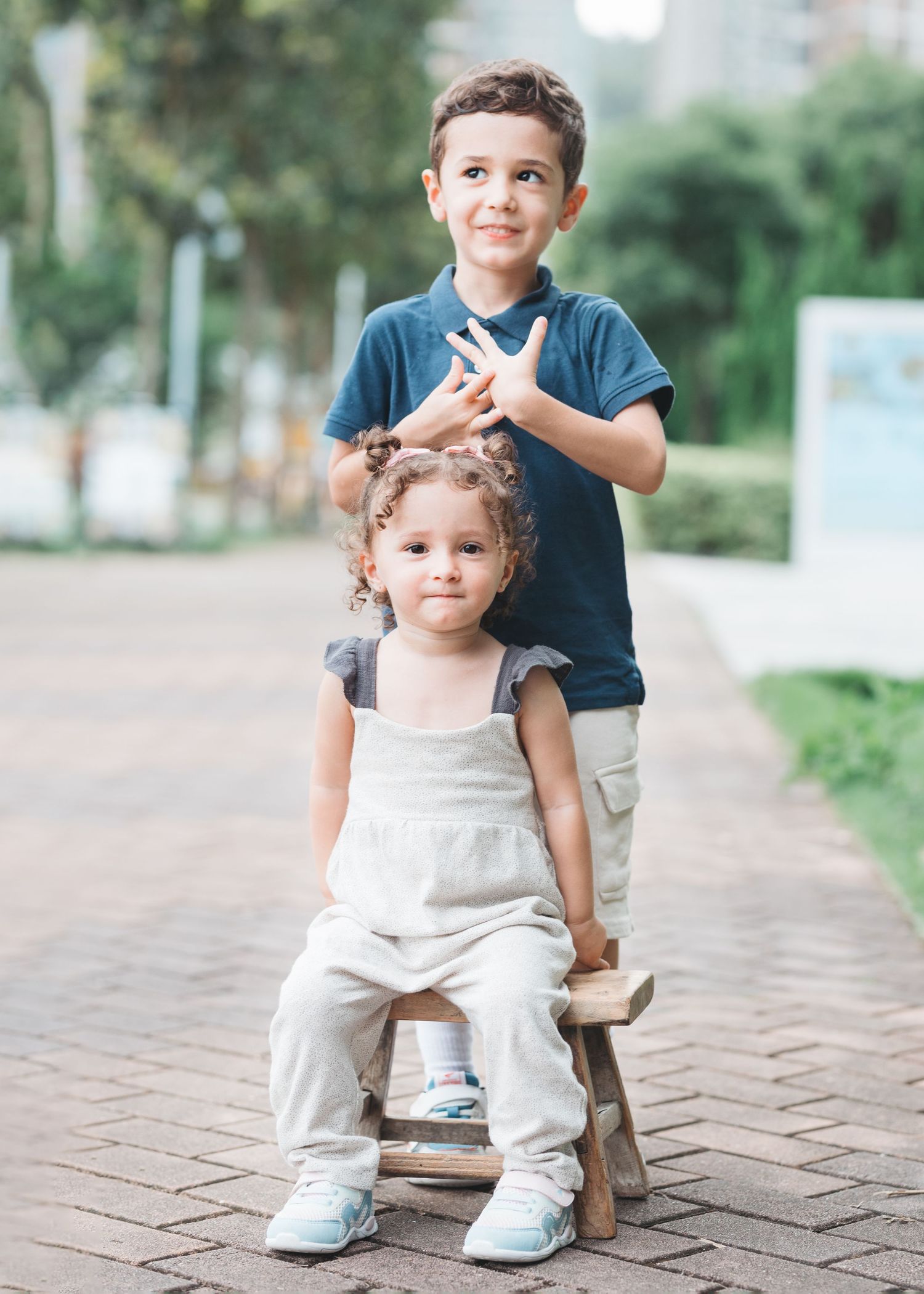 Sweet toddler in beige overalls and white sneakers creates playful poses on an outdoor brick walkway. Family Photographer in Hong Kong